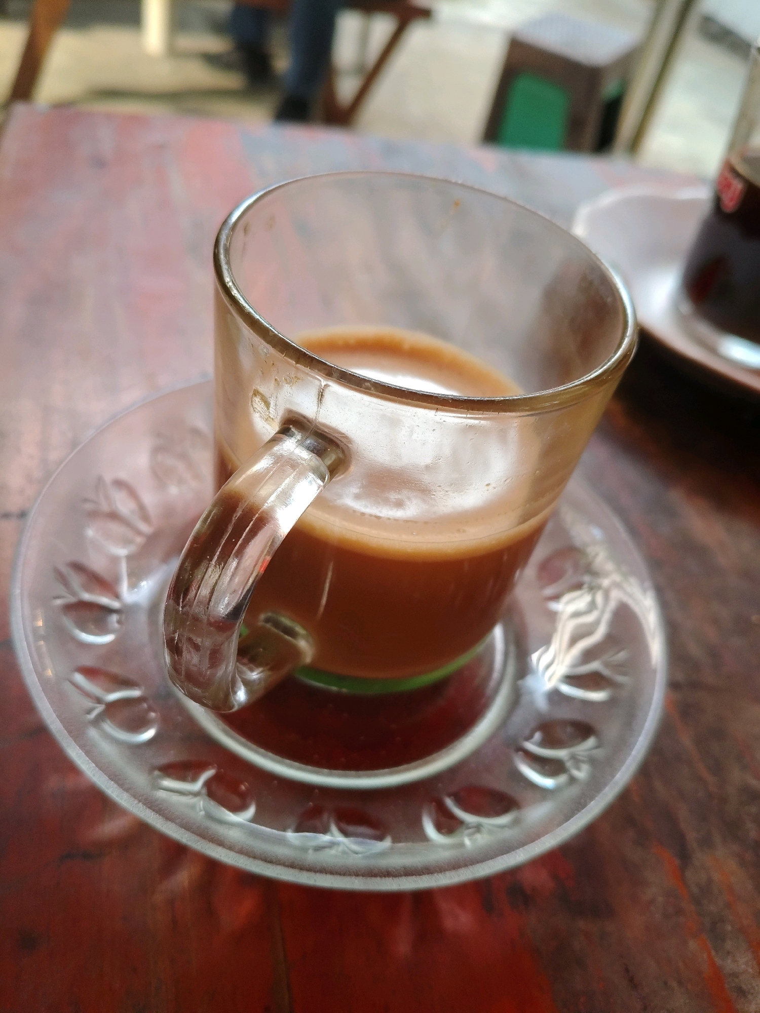 A close-up, eye-level shot shows a clear glass mug filled with a light brown beverage, likely coffee or tea with milk, resting on a clear glass saucer with a decorative floral pattern. The mug is positioned on a reddish-brown wooden table with a visible grain pattern. A second, out-of-focus white saucer with a dark beverage is visible to the right. In the blurred background, a person in blue jeans and a dark shirt can be seen standing, with parts of wooden furniture and a green object suggesting an indoor or semi-outdoor setting, possibly a cafe. The overall lighting is soft and natural, with a shallow depth of field that emphasizes the mug and saucer. The scene is set in South Jakarta, Indonesia.