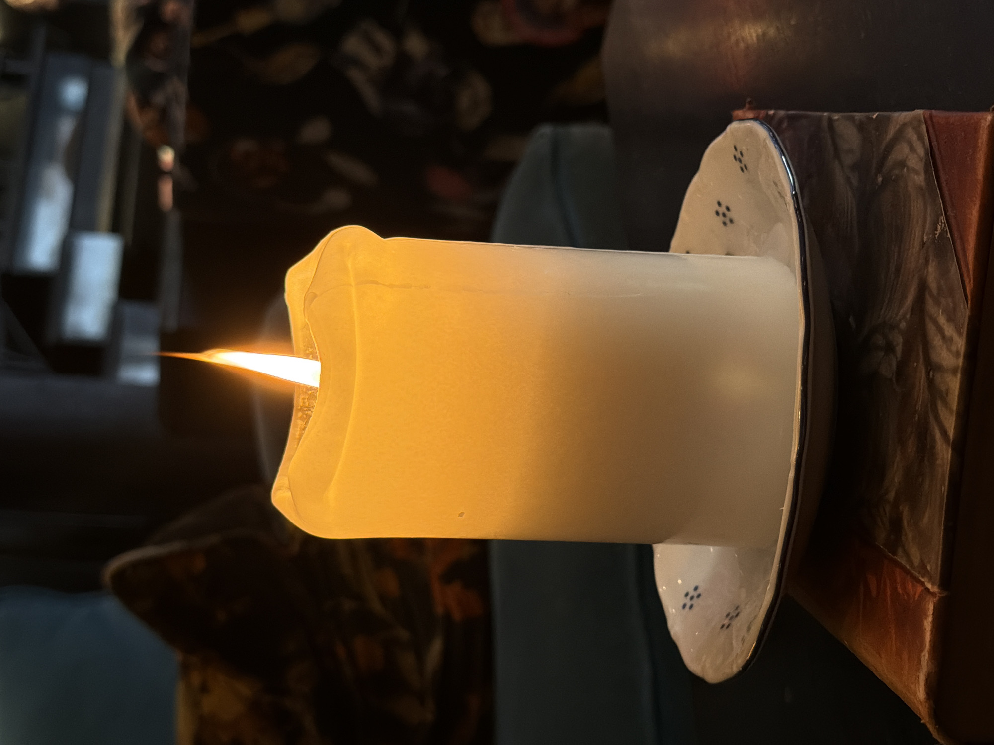 A lit, thick white pillar candle with an orange flame is positioned on a white saucer with blue markings. The saucer rests on a closed book with a reddish-brown cover. The background is dark and out of focus, with hints of patterned upholstery and furniture. The scene appears to be indoors, possibly in a dimly lit room. There are no people visible. The image is a still life focusing on the candle and its immediate surroundings. The location is not discernible from the image.