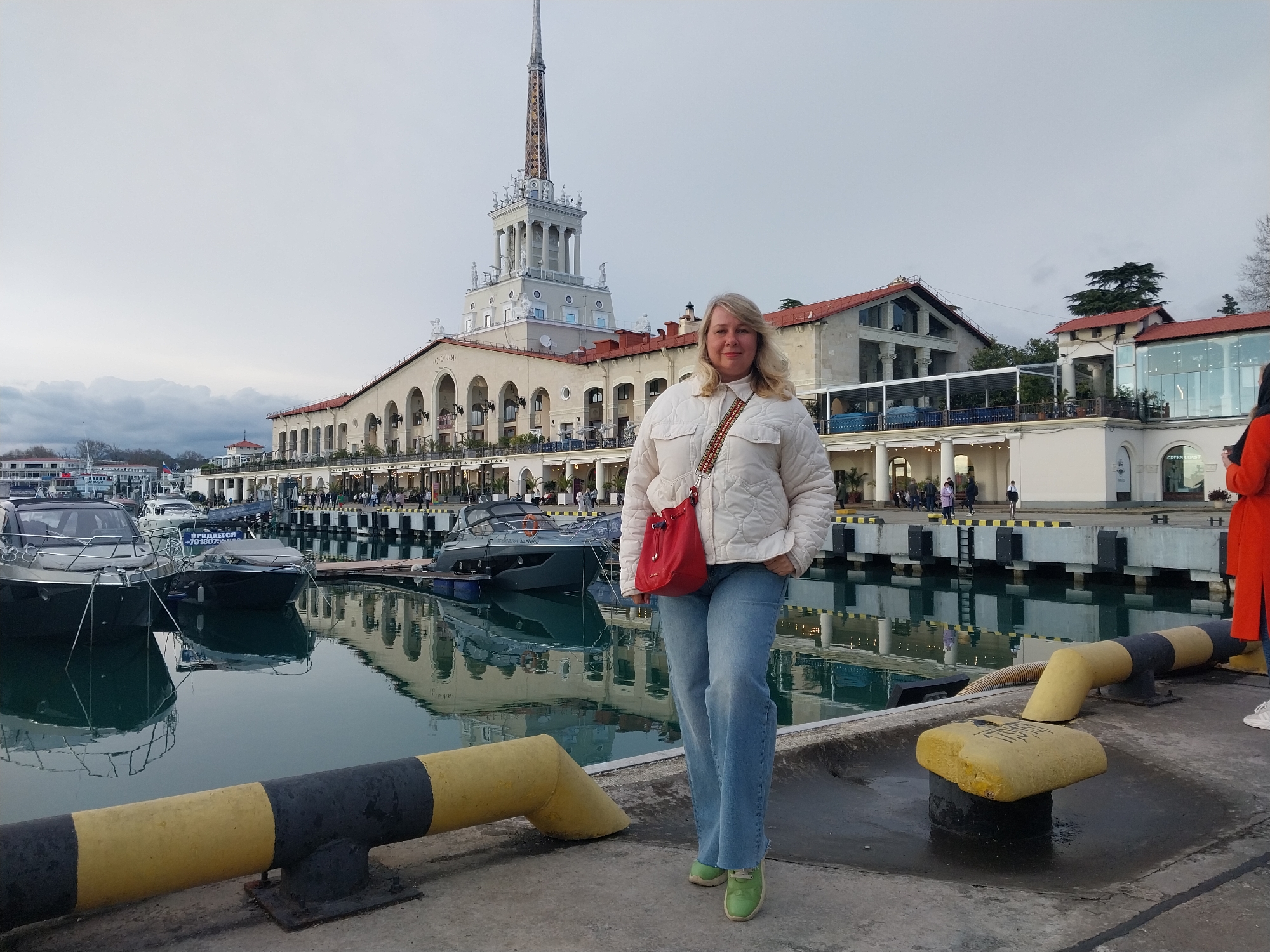 A woman stands on a pier in Sochi, Russia, with the Sochi Marine Station visible in the background. The sky is overcast.
The woman, who appears to be middle-aged, is wearing a white jacket, light blue jeans, and green shoes. She has a red shoulder bag. Her hands are in her pockets. She is the central subject of the image.
Behind her, the Sochi Marine Station dominates the scene with its iconic tower and grand architecture. The building's facade is reflected in the calm waters of the harbor. Several boats are docked in the harbor. Yellow and black bollards line the edge of the pier.
People are walking in the background along the colonnade of the Marine Station. Another person, wearing an orange coat, is partially visible on the right edge of the frame.