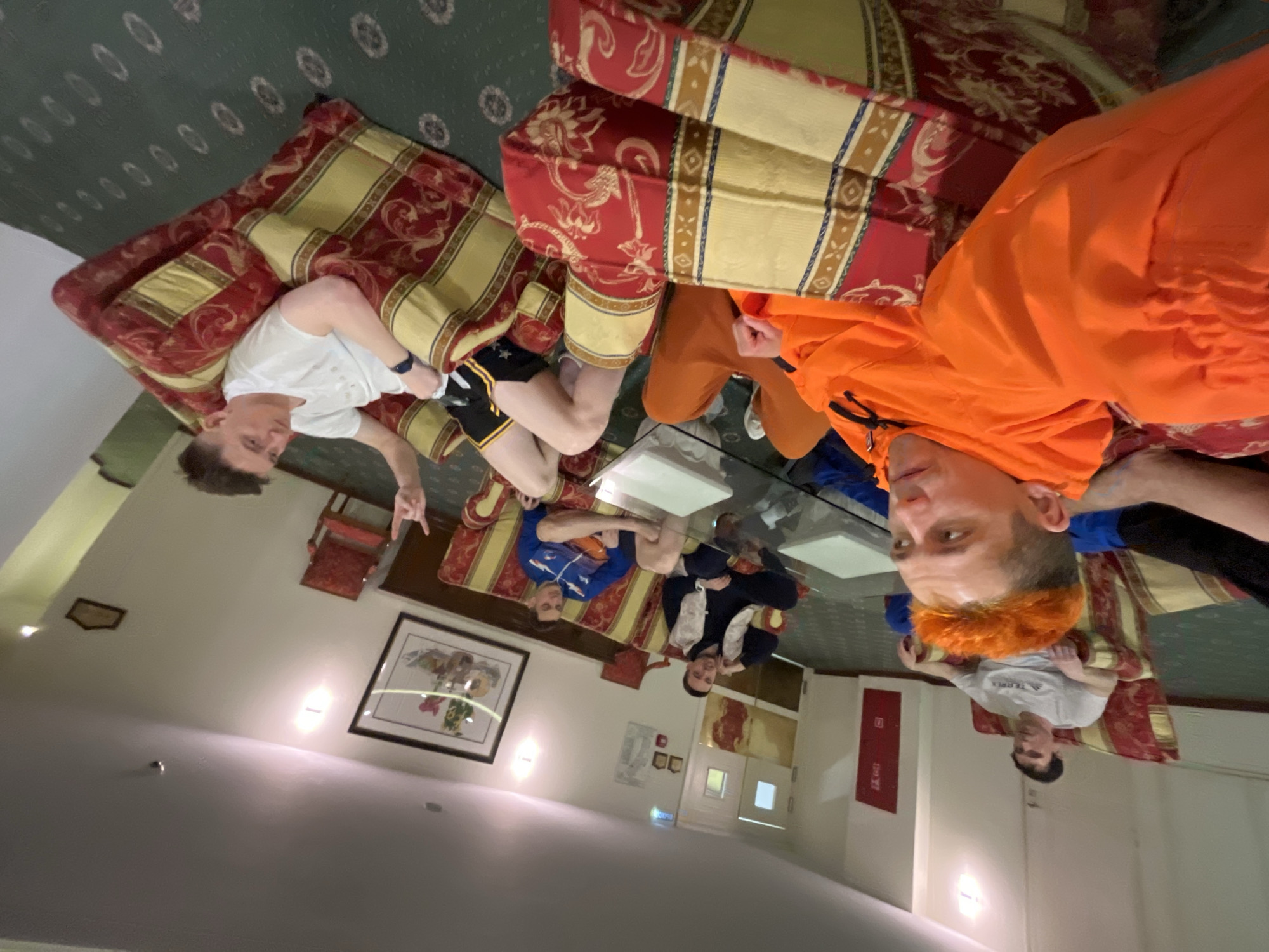 A group of six people are seated on red and gold couches around a glass-topped coffee table. A man in a white t-shirt and black shorts is lying on a couch, giving a peace sign. A man in an orange shirt is leaning back on the couch, facing the camera. Two men in blue shirts are seated next to each other. Another man in a dark suit is seated next to them. A man in a grey t-shirt is seated on a couch near a fire extinguisher. The setting appears to be a hotel lobby or lounge. A painting is hanging on the wall, and there are signs in Russian visible in the background, suggesting the location is in Moscow, Russia.