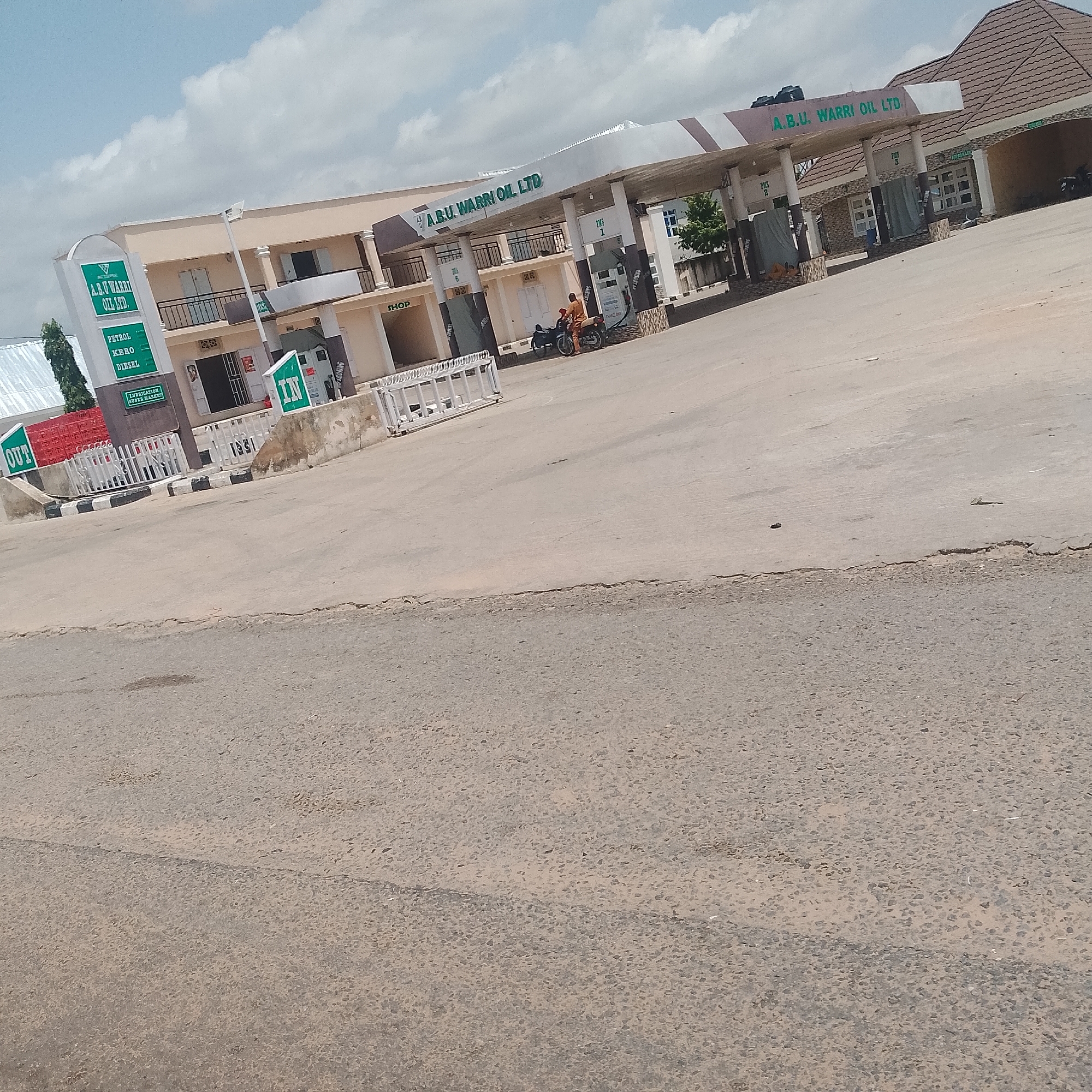 The image shows a street-level view of a gas station in Jalingo, Nigeria. The gas station is identified by signage as "A.B.U. WARRI OIL LTD." The station features a covered canopy over multiple fuel pumps, some of which are covered with tarps. On the left side of the station, there are green signs indicating "OUT" and "PETROL KERO DIESEL". Another green sign with "IN" is visible on the right side. A shop area is also present within the station complex. A person on a motorcycle is stopped near one of the fuel pumps. The ground in front of the station is paved, and the area beyond the paved surface is an asphalt road. The sky is partly cloudy. The overall environment suggests a typical urban or semi-urban setting.