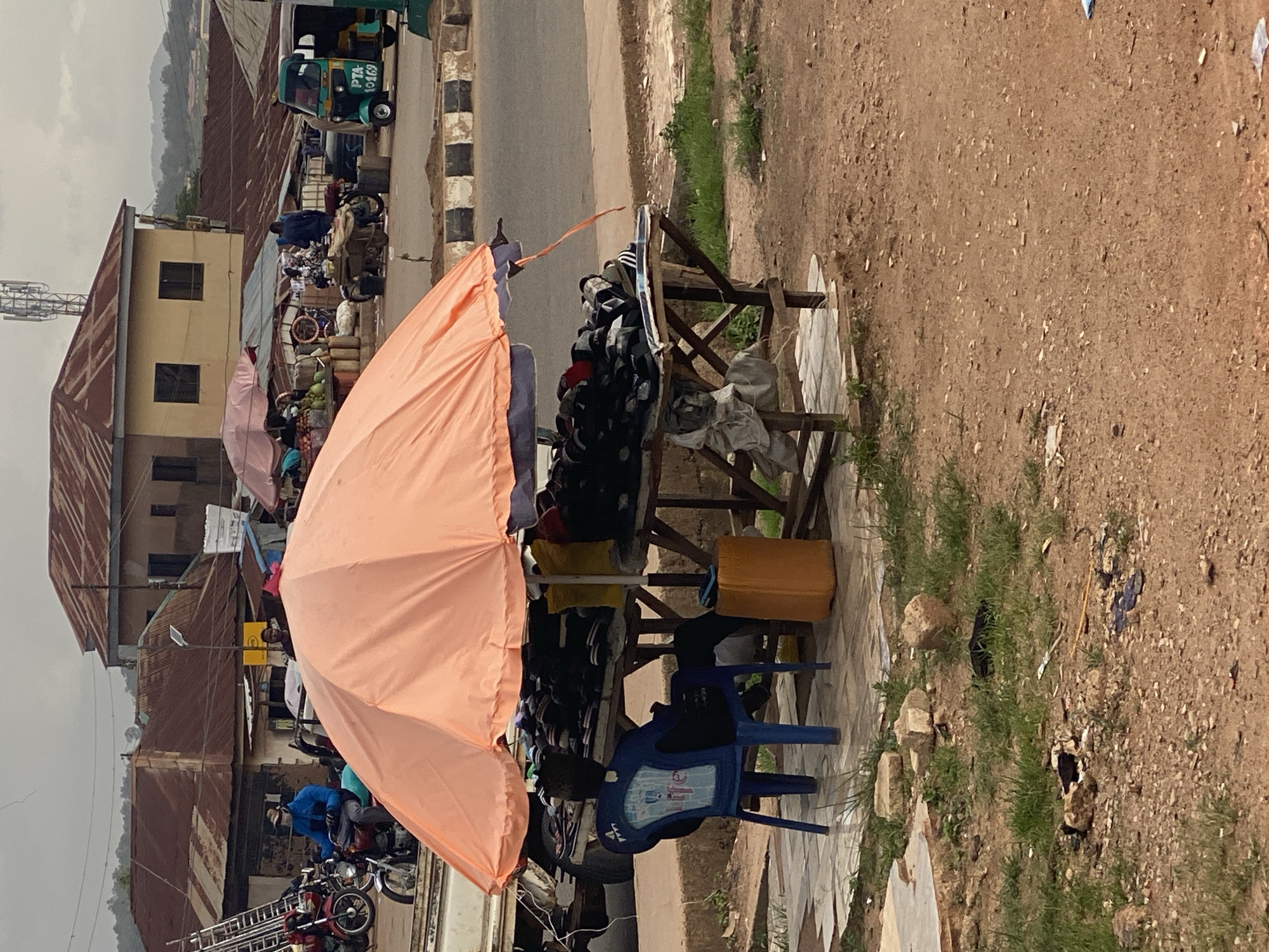 The image depicts an outdoor market scene in Gura Top, Nigeria. A stall, shaded by a large, pale orange umbrella, displays various items, including what appear to be hats and other clothing accessories arranged on wooden shelves. A blue plastic chair sits in front of the stall. Behind the stall, a dirt path or ground is visible, with sparse green grass and scattered rocks.
In the background, buildings with corrugated iron roofs line a street. A green and white tricycle, possibly a form of local transport, is parked on the roadside, with the license plate "PTA 10469" visible. Further down the street, more stalls and a collection of motorcycles suggest a bustling marketplace.
A person wearing a blue jacket is visible near some motorcycles, interacting with them. The overall scene suggests a daytime setting, likely mid-morning or afternoon given the lighting. The weather appears to be overcast, as indicated by the diffused light and the muted colors of the sky in the background. The atmosphere seems ordinary, depicting a typical day in a Nigerian market. There are no overt emotional expressions visible in the people, and the focus is on the commercial activity and the environment.
