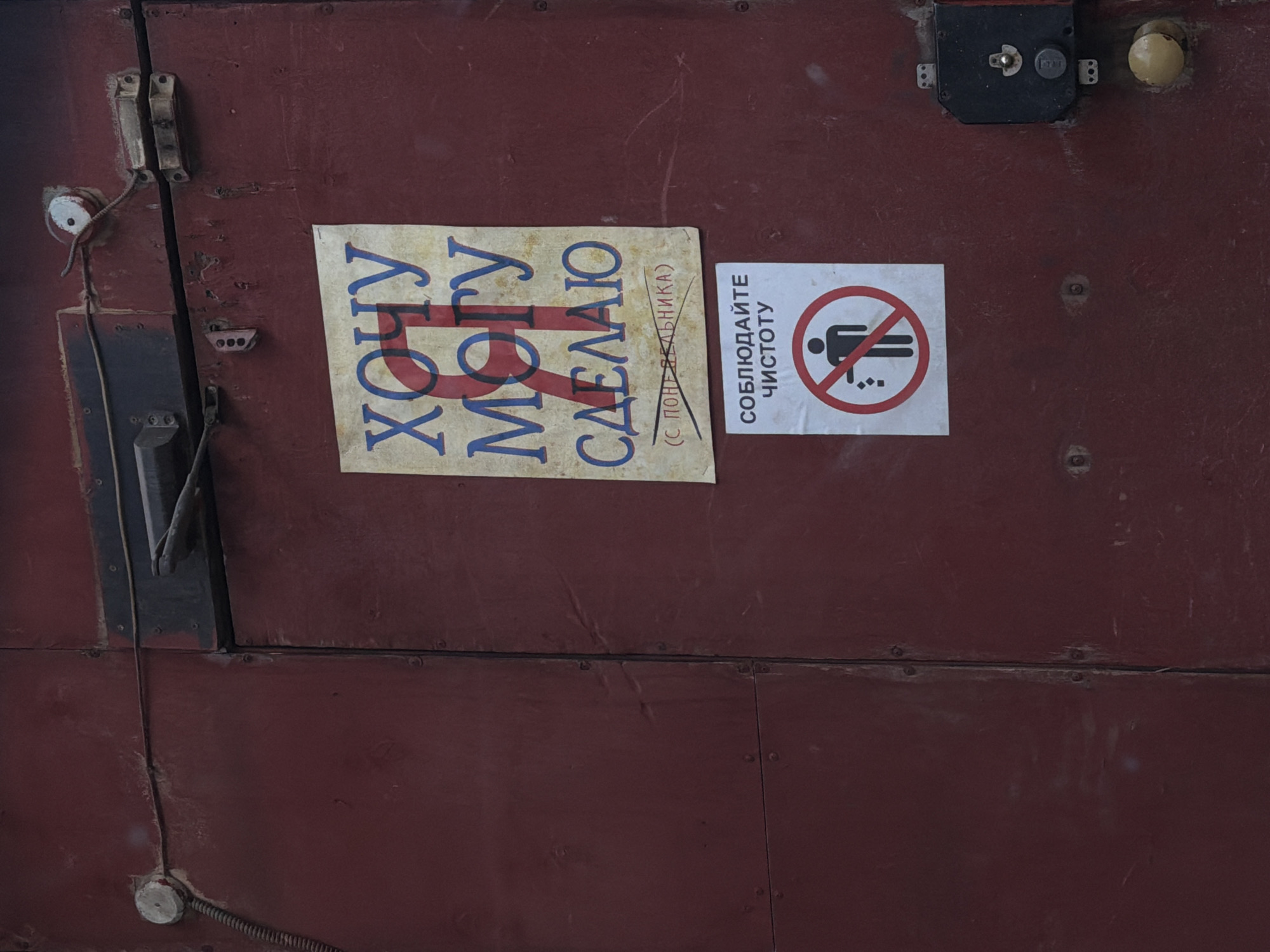 A close-up shot features a red metal door or wall with various fixtures and signs. On the left side is a metal panel with a handle and what appears to be electrical wiring and components. There are two signs affixed to the surface. The first sign, to the left of the center, contains text in Cyrillic script in a blue font, and the text appears to be superimposed with a red line. To the right of the first sign is a white square sign with a red circle and a line through it, depicting a stick figure in a crossed-out position. The sign also contains Cyrillic text, possibly instructions or warnings. At the top right corner of the frame, there is a black lock and a yellow doorknob. The metal surface exhibits signs of wear and tear, including scratches and small dents. The location is unknown, as there is no information present to identify the city and country.