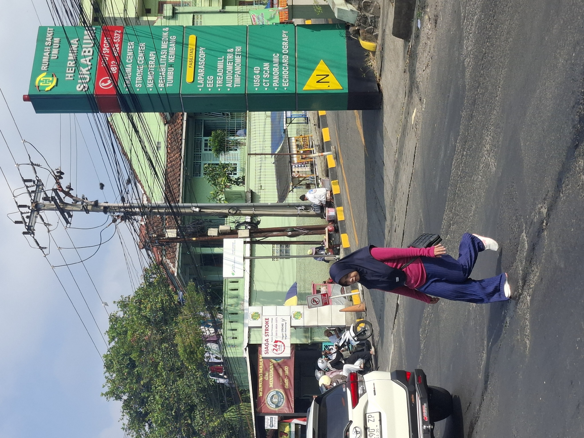 The image depicts a street scene in Sukaraja, Indonesia, featuring a tall green hospital sign for "Rumah Sakit Umum Hermina Sukabumi" prominently displayed on the right. The sign lists various medical services, including "Trauma Center," "Stroke Center," "Kemoterapi," and "Rehabilitasi Medik & Tumbuh Kembang," as well as facilities like "Laparoscopy," "EEG," "Treadmill," "Audiometri & Timpanometri," "USG 4D," "CT Scan," "Panoramic," and "Echocardiography." The hospital building, painted in a light green hue, is visible in the background.
In the foreground, a woman wearing a dark blue hijab and a magenta long-sleeved shirt, paired with loose blue trousers, walks across the asphalt road. She is carrying a black shoulder bag. To the left, a white SUV is parked, and further back, several people on motorcycles are stopped. A red and white banner for "Warung Makan dan Minuman" is visible on the left side of the street. The environment includes trees, power lines crisscrossing overhead, and a clear blue sky. The road surface shows signs of wear and tear.