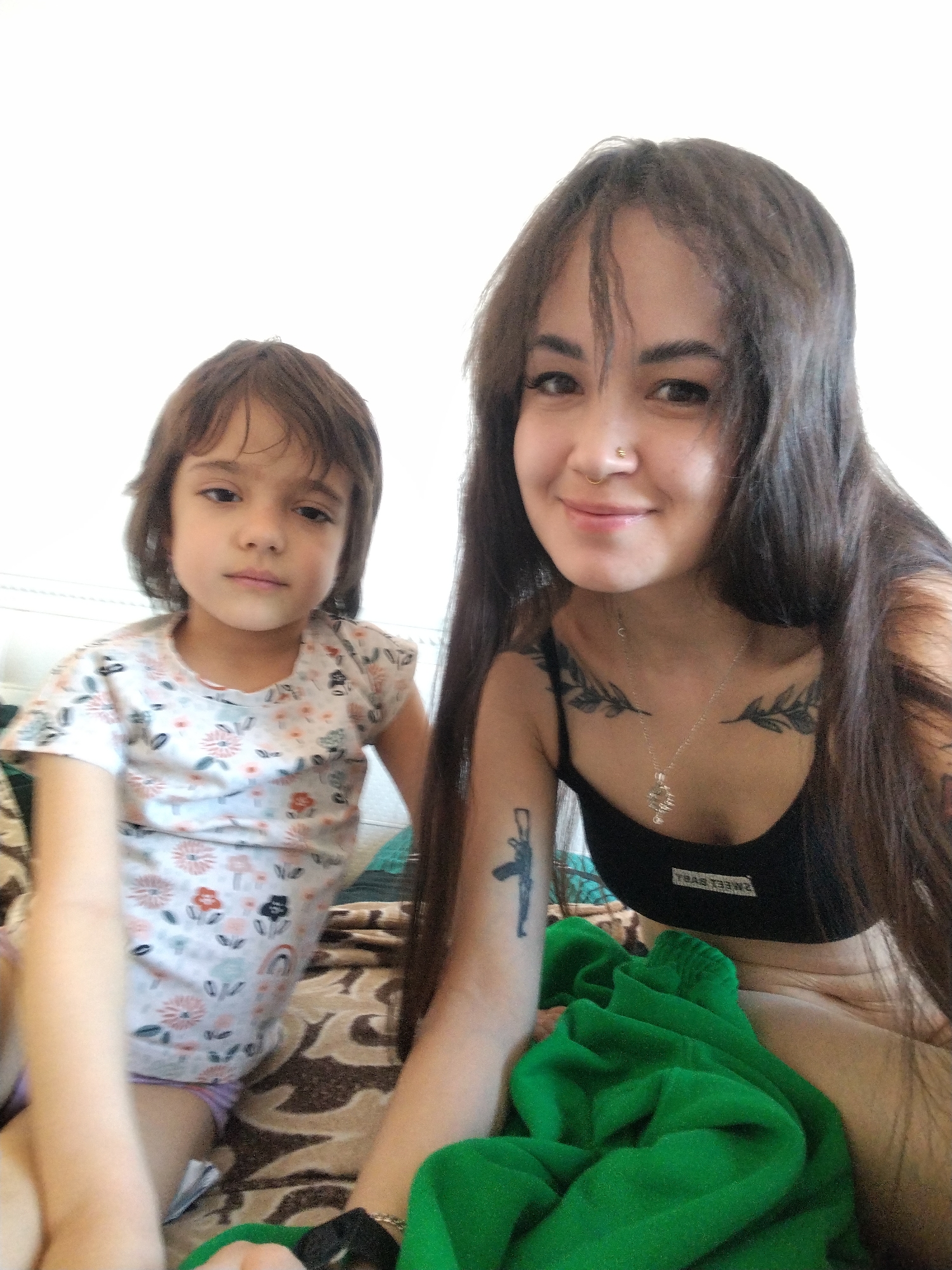 In an indoor setting in the city of Unknown, Unknown, a woman and a young child are seated close together. The woman, with long dark hair and a nose ring, is wearing a black top and a necklace, displaying tattoos on her chest and arm. The child, with short brown hair and a light complexion, is wearing a patterned t-shirt. A green garment is draped nearby, and the two are sitting on a patterned surface, possibly a bed or couch.