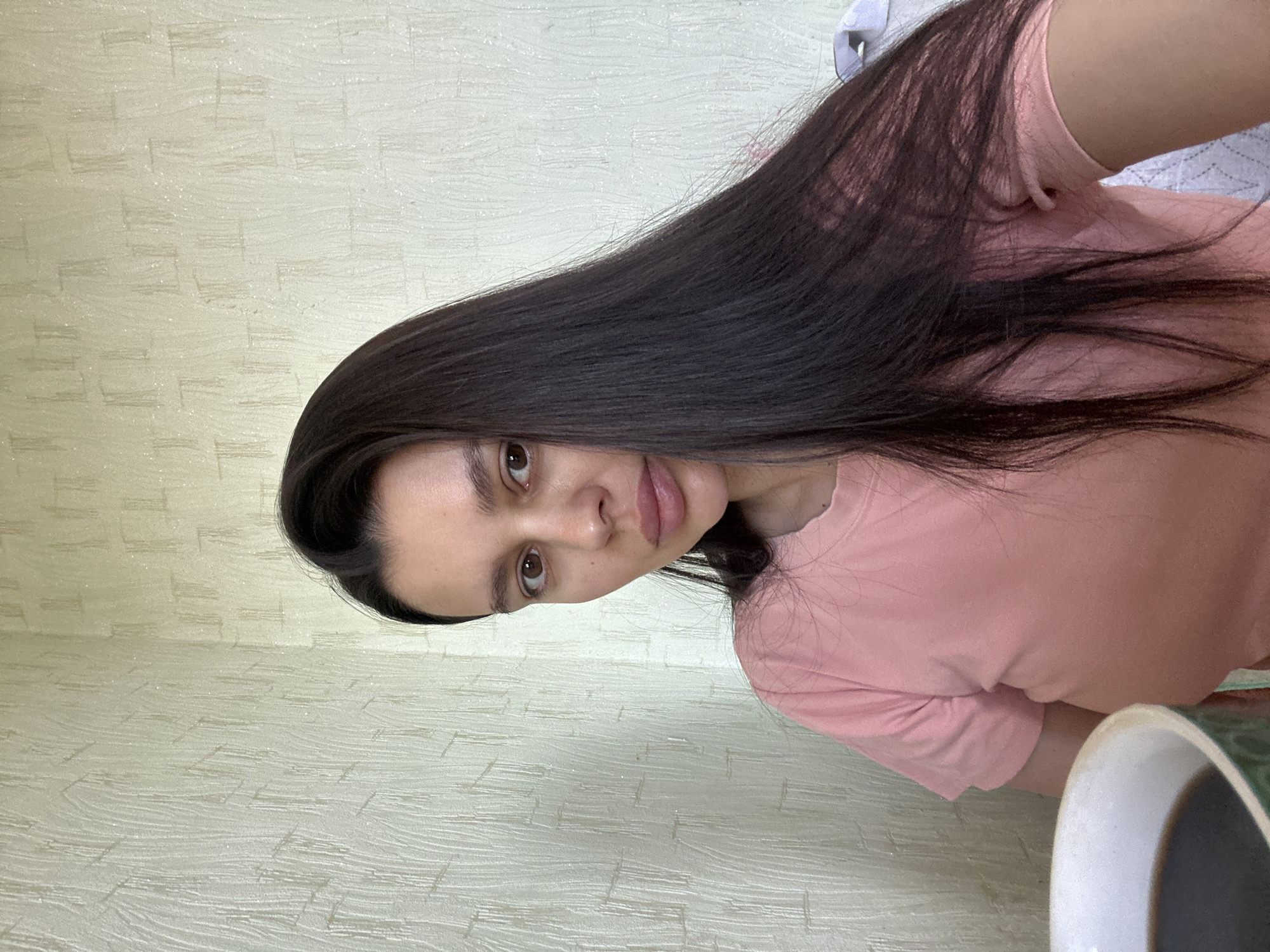 A woman with dark hair is wearing a pink shirt and looking at the camera. The background is a textured, light-colored wall. A cup is visible in the bottom right corner. The location is not discernible from the image.