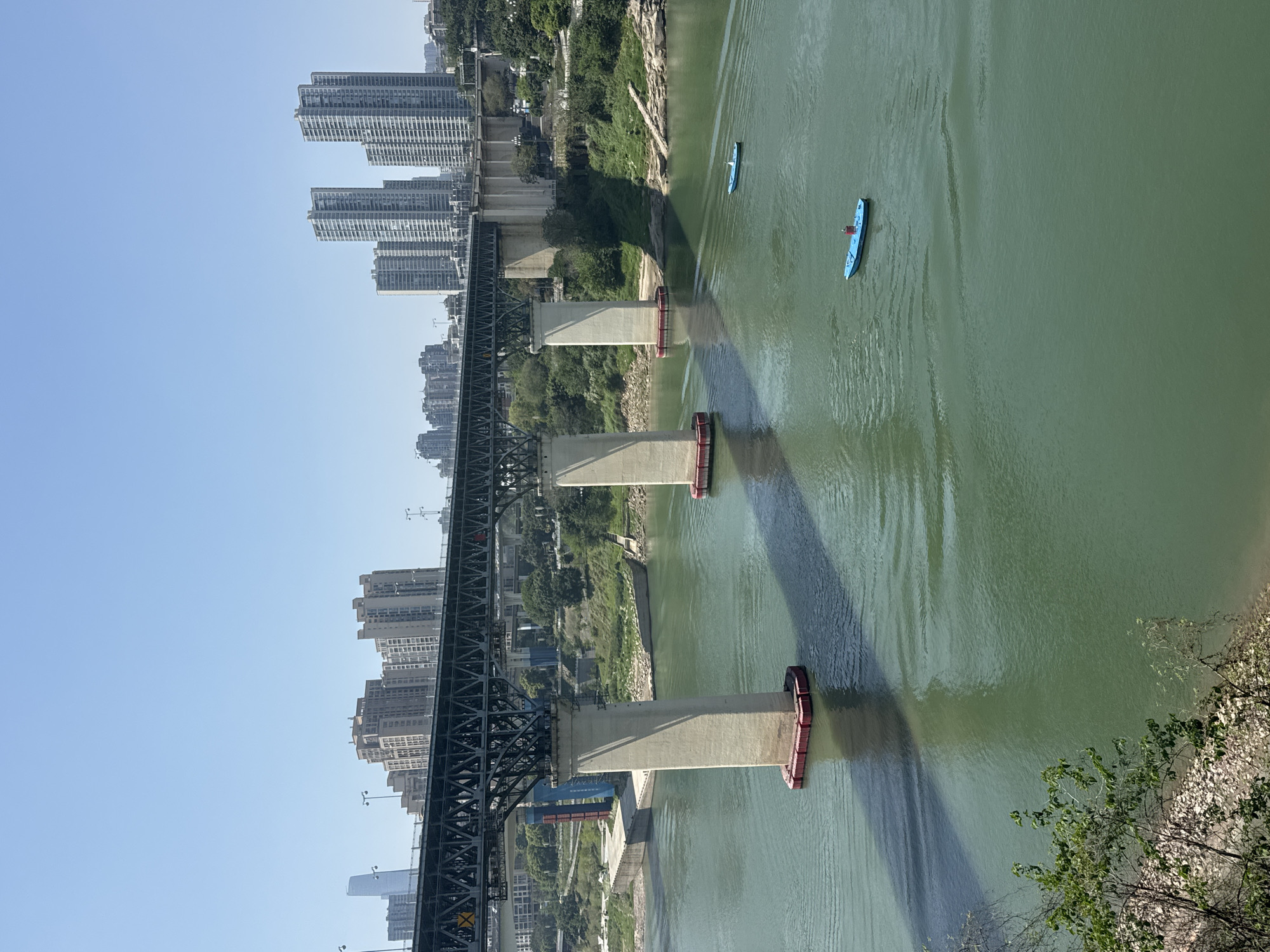 The image shows a view from a bridge in Yuzhong District, China, looking down at a river. A steel truss bridge crosses the river. Large concrete support pillars are visible in the water, casting shadows. Two blue kayaks are on the water, with one being paddled. High-rise buildings are on the riverbank near the bridge. There are vegetated slopes to the side of the bridge. A rocky bank with shrubs can be seen in the foreground. The sky is blue and clear.