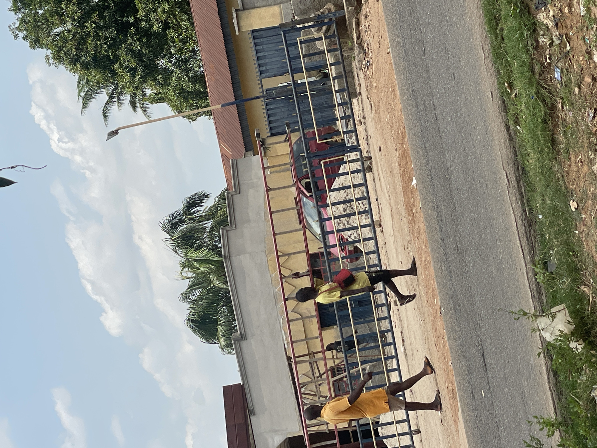 The image, rotated 90 degrees clockwise from its correct orientation, depicts a bright, sunny daytime scene in what appears to be Mararaba, Nigeria, featuring a roadside area.
In the foreground, a dirt path runs parallel to a narrow, paved road. A low metal fence, painted with red and blue sections, separates this path from a property behind it. Two young individuals are visible near this fence, both wearing yellow t-shirts and shorts. One person in the immediate foreground wears light-colored shorts and stands barefoot or in sandals. Further back, another person wears dark shorts, has a red bag slung over their shoulder, and also appears to be barefoot or in sandals. Their activity is not clearly discernible; they seem to be standing or walking along the fence.
Behind the fence, a light-colored building with a dark red corrugated iron roof is visible, along with a large, dark blue metal gate. A red car is partially visible behind this gate, parked within the property. Lush green trees and palm fronds are abundant in the background and above the structures, indicating a tropical climate. A street light pole extends into the upper part of the image (when correctly oriented), with its lamp fixture positioned over the road.
The sky is clear blue with scattered white clouds, and the strong sunlight casts clear shadows, confirming it's daytime. The roadside features a dirt shoulder with some green weeds and scattered debris. No discernible text is visible in the image.