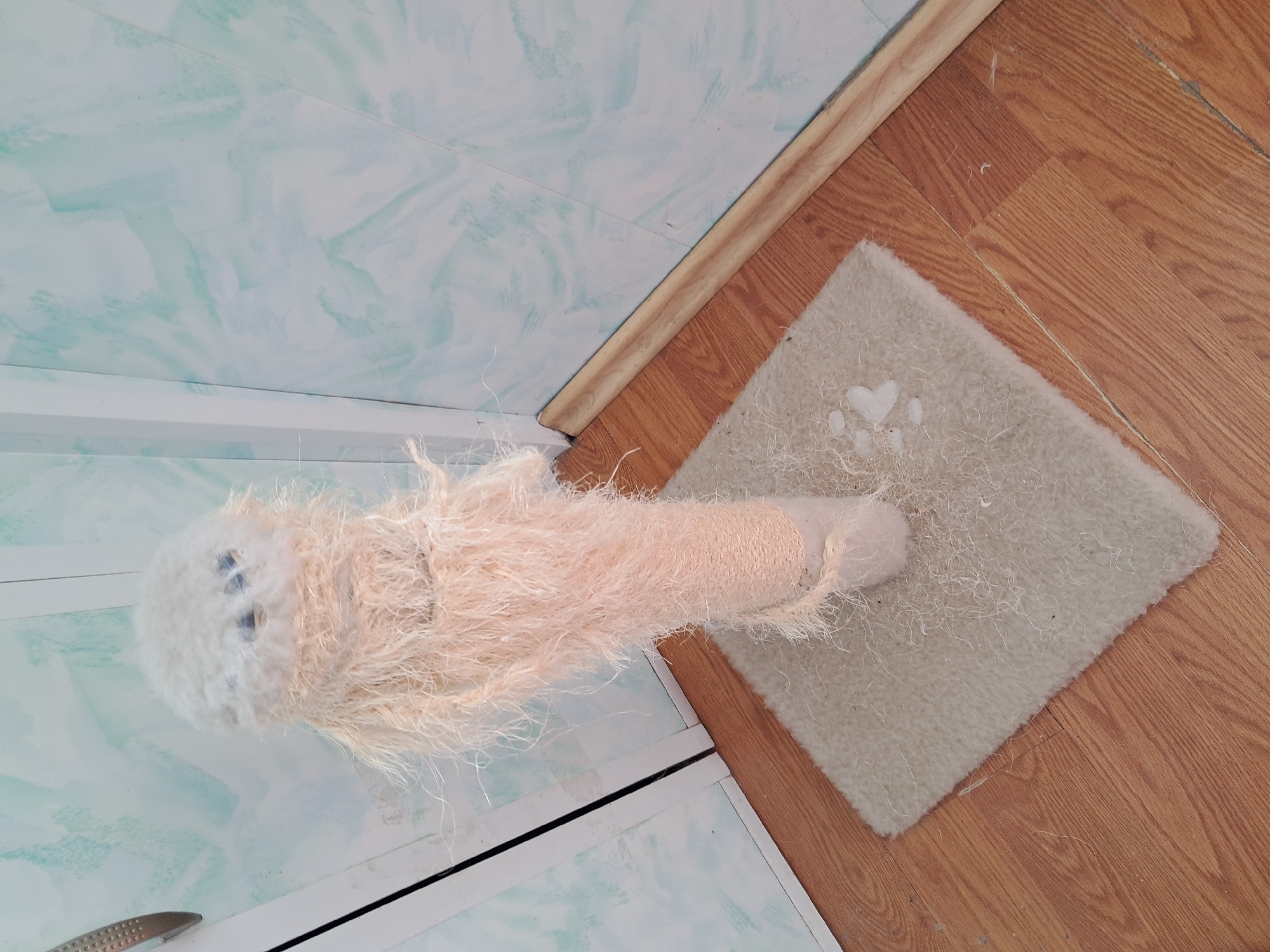 An angled, high-view indoor shot captures a well-used cat scratching post and an accompanying mat on a wooden floor, set against a patterned wall in Kyiv, Ukraine. The dominant subject is a vertical cat scratching post, topped with a fluffy, light cream or off-white spherical element that features two small dark indentations, possibly decorative or due to wear. The post's main body is wrapped in a light peach-colored, long-pile fluffy yarn, transitioning to a section of natural fiber rope near its base. Numerous loose fibers are visible on both the fluffy yarn and the rope, indicating significant use.
The scratching post stands on a small, rectangular mat, light beige or off-white in color, with a slightly shaggy texture. Several white paw print shapes are visible on the mat's surface. This mat rests on a floor composed of light brown wooden planks with distinct grain patterns.
A light-colored wooden trim separates the wooden floor from the wall. The wall itself features a light blue-green background with a subtle, lighter marbled or swirled pattern. No people or animals are present, and no discernible text is visible. The scene suggests a home environment, with objects showing signs of regular pet activity.