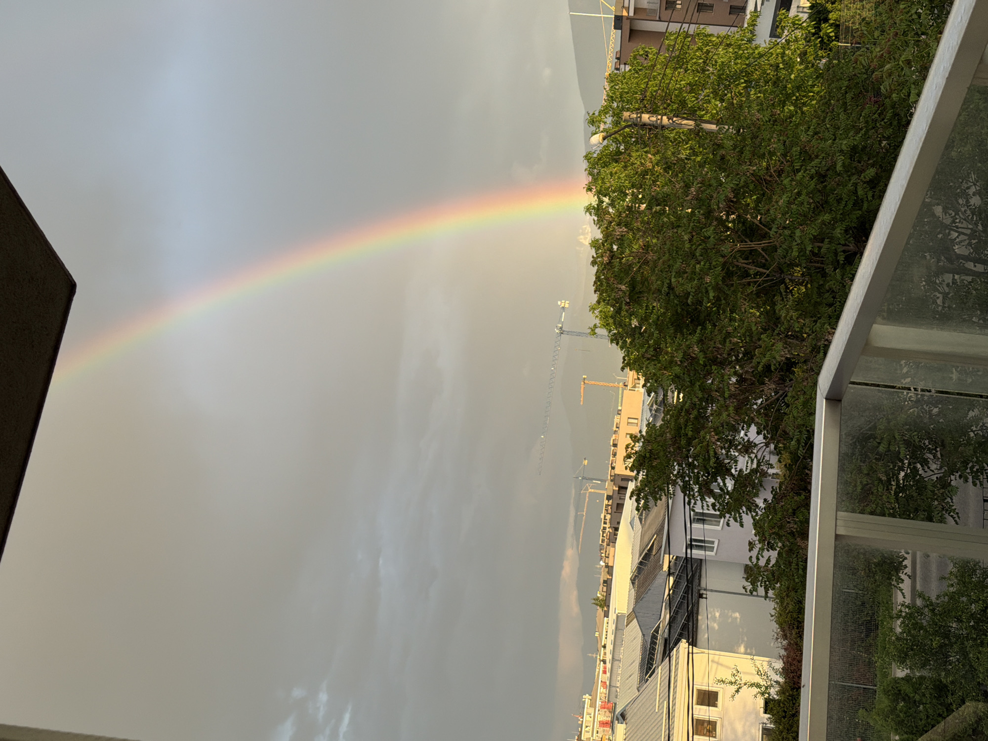 The image shows a rainbow in the sky over Plovdiv, Bulgaria. The rainbow stretches from left to right, arching over a cityscape with buildings, trees, and construction cranes. The sky is cloudy and gray. The photo was likely taken from a balcony or elevated position, with a railing visible in the foreground.
