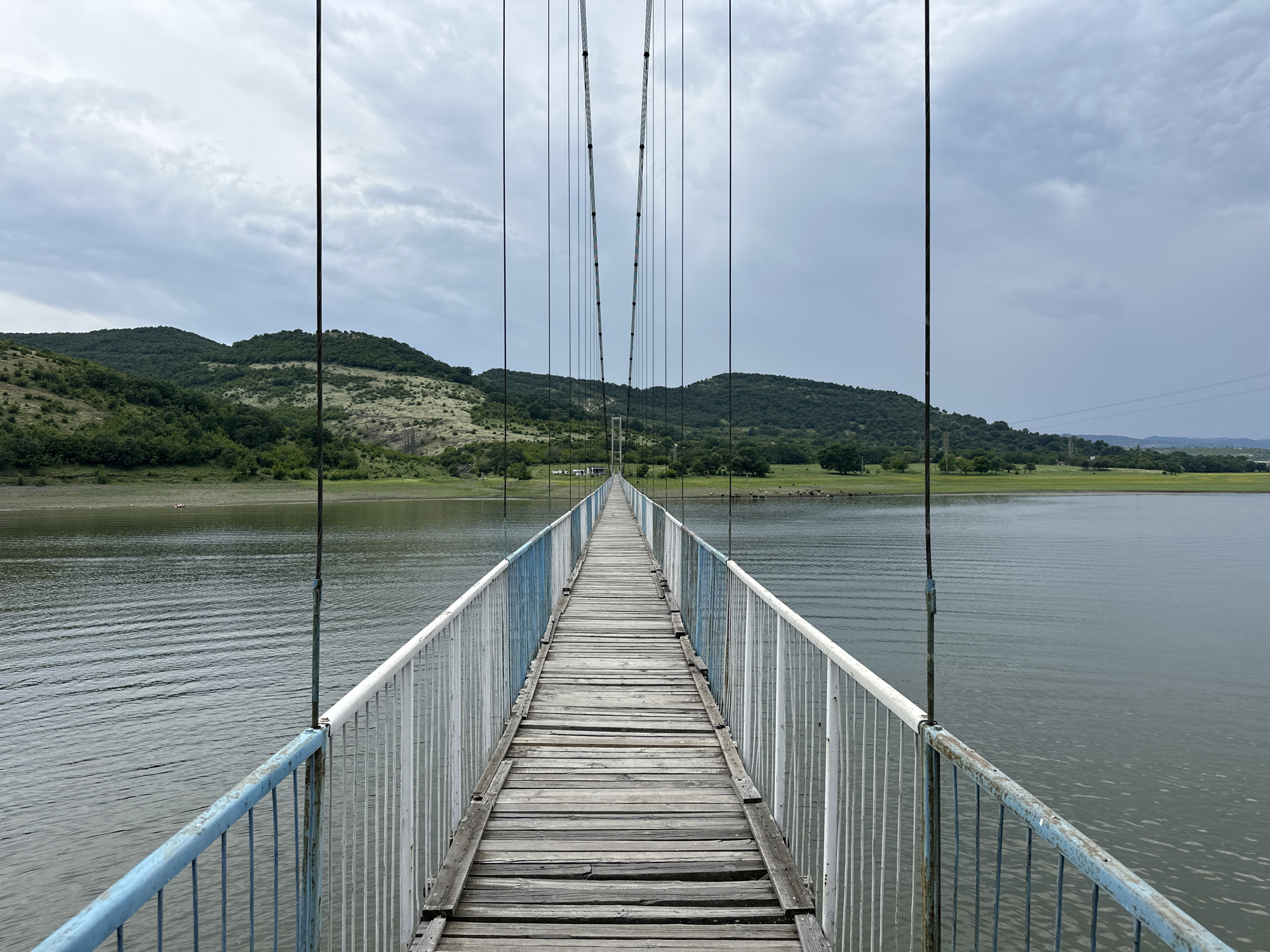 The image presents a long, narrow suspension bridge extending across a body of water in Lisitsite, Bulgaria, viewed from a pedestrian perspective looking straight down its length. The bridge features a walkway constructed from weathered, light-colored wooden planks, many showing signs of age and wear. Along both sides of the walkway are metal railings, painted in alternating sections of faded blue and peeling white. Slender black suspension cables descend vertically from above, connecting to the main support cables which converge towards a distant, smaller suspension tower at the far end of the bridge.
The water beneath the bridge is a calm, dark greenish-grey, with subtle ripples visible across its surface. Beyond the water, on both sides, rise undulating hills densely covered with a mix of green trees and lighter patches of sparse vegetation. The sky above is overcast, filled with soft, grey clouds. At the distant end of the bridge, on the far bank, a small white building and several vehicles are visible. Further in the background, a flat plain extends, featuring scattered trees, and distant utility poles with power lines are discernible on the right side. No people are present on the bridge or in the immediate surroundings.