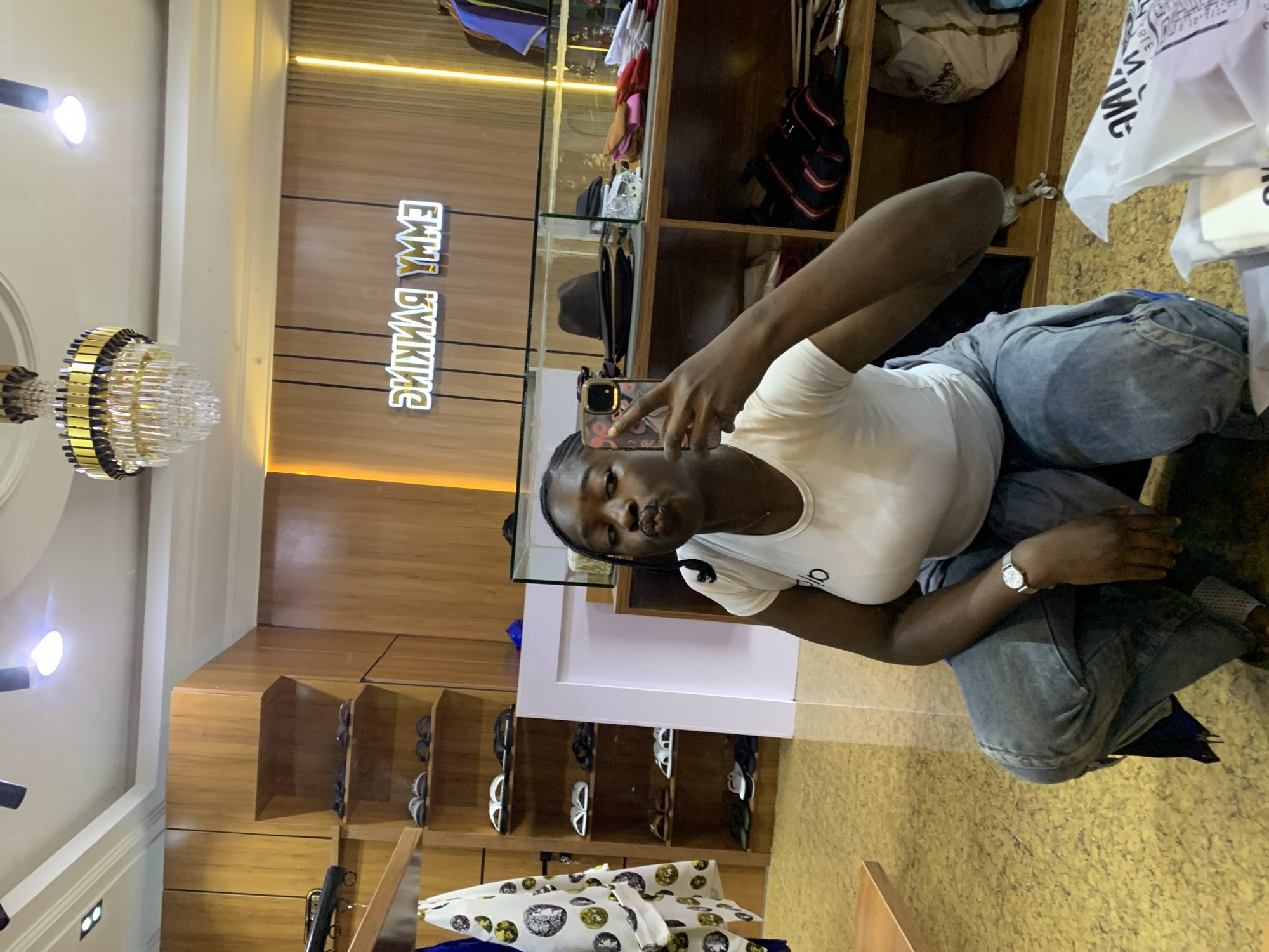 The image depicts a young Black woman taking a mirror selfie in what appears to be a boutique or clothing store. She is squatting down, wearing a white t-shirt and loose-fitting denim pants. She has her phone held up to take the photo, and her lips are puckered. The background shows wooden shelves stocked with various items, including shoes, hats, and clothing. A neon sign spelling "EMMY RANKING" is visible on the back wall. The store environment suggests it could be located in Gura Top, Nigeria, given the context. The lighting is bright, with a chandelier visible on the ceiling.