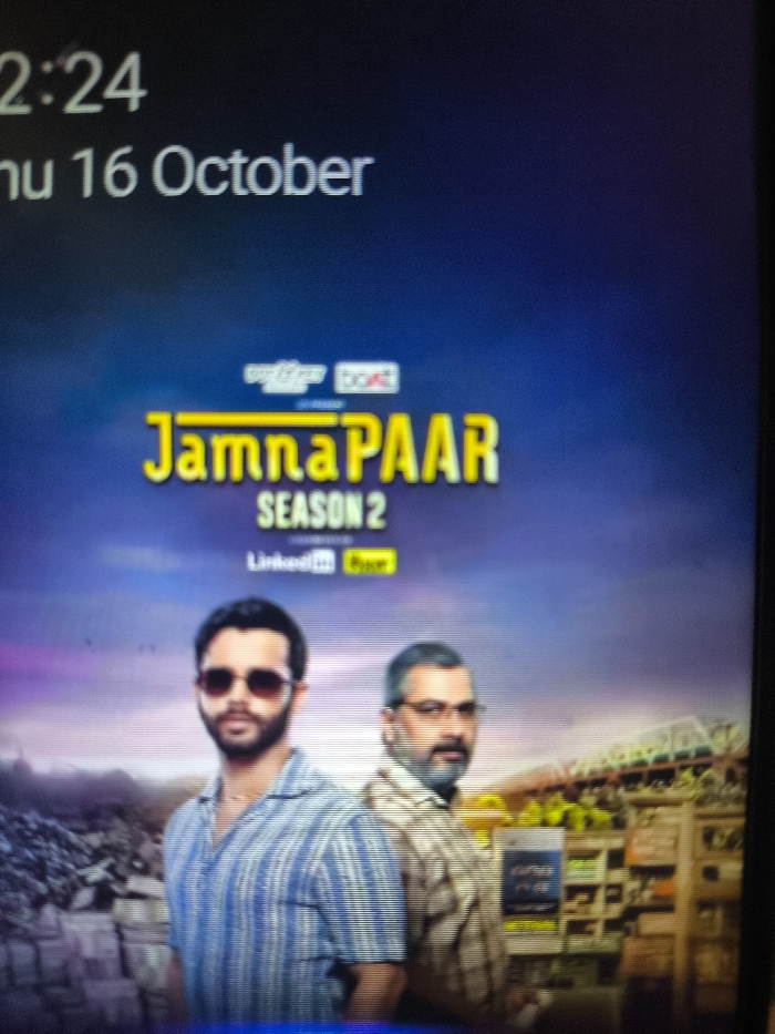 The image depicts a photograph of a digital screen displaying a promotional poster for "Jamna PAAR Season 2." In the upper left corner, the time "2:24" and date "Thu 16 October" are visible. The poster features two men in the foreground: the individual on the left has a dark beard, wears sunglasses, and a blue and white striped collared shirt, facing forward; the individual on the right has grey hair and beard, wears spectacles, and a brown checkered shirt, turned slightly towards the left of the frame. Above the "Jamna PAAR" title, the logos "myfnp." and "boAt" are present. Below "SEASON 2," the logos "LinkedIn" and "Cheez" are displayed. The background behind the figures is a stylized urban landscape, featuring what appears to be a debris-strewn area on the left and a cluster of buildings with a large stadium-like structure in the distance on the right, all under a sky transitioning from purple at the horizon to dark blue at the top. The image contains no visual elements or discernible landmarks that identify a specific location within Nicosia, Northern Cyprus / Cyprus.