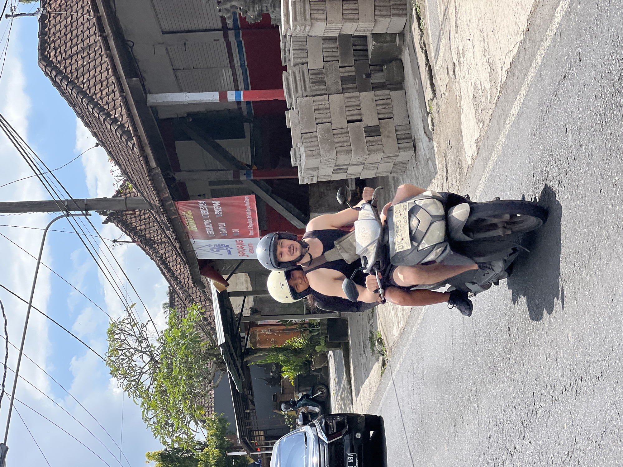 In the city of Ubud District, Indonesia, two individuals are riding on a scooter. The person in front, a man, is wearing a black tank top, black shorts, black sneakers, and a dark grey helmet. He is operating the scooter. The person behind, a woman, is wearing a black shirt and a white helmet. They are both facing forward and appear to be traveling on a paved road. To the left of the scooter, a black car is visible on the same road. The background consists of buildings with tiled roofs, some vegetation, and utility poles with numerous wires crisscrossing overhead. The sky is blue with scattered clouds. To the right of the road, stacks of concrete blocks are piled up. A red banner with text in Indonesian is hanging from one of the buildings.