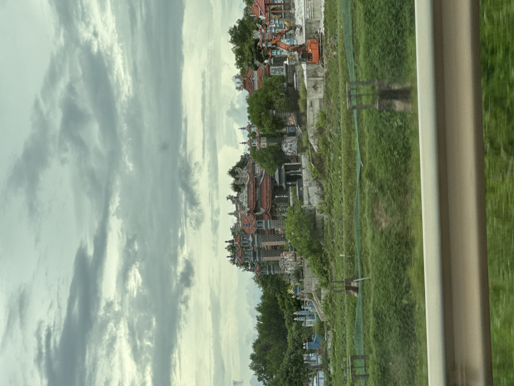 The image shows a cemetery in Hải Lăng Commune, Vietnam. The sky is overcast with heavy, gray clouds. In the foreground, there is a grassy area with some fencing and a concrete barrier. Behind this, the cemetery is visible with its elaborate tombs, some of which have red tiled roofs and ornate decorations. Several trees and lush green vegetation surround the tombs. An orange excavator is parked to the right of the cemetery, suggesting ongoing construction or maintenance. The overall scene is somber, characterized by the cloudy sky and the presence of a cemetery.