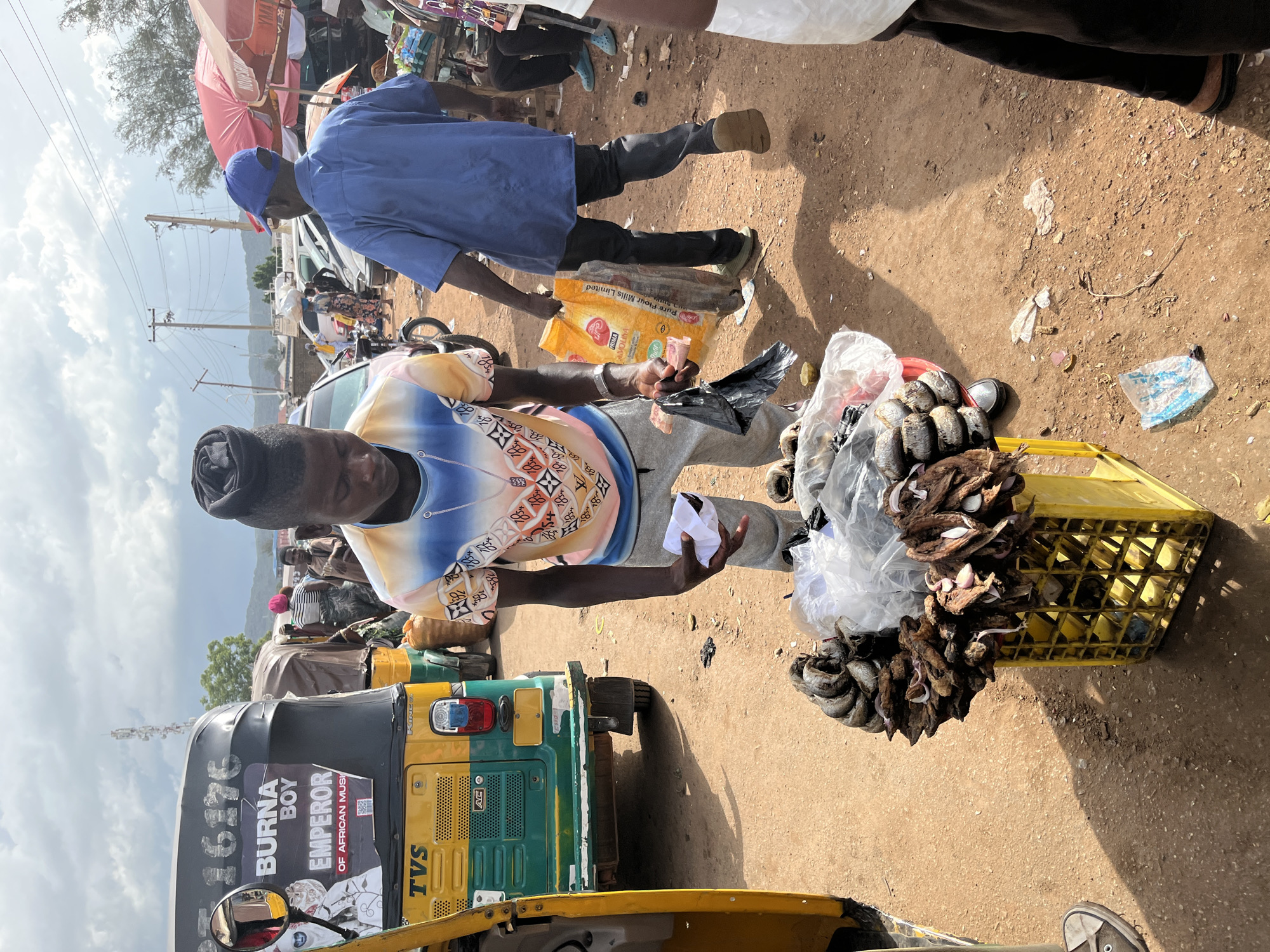An outdoor market scene is depicted in Rafin Par, Nigeria, under a cloudy sky. The central figure, a young man wearing a colorful patterned t-shirt and grey sweatpants, stands next to a yellow crate filled with dried fish and onions. He is holding a white paper in his right hand and a black plastic bag in his left. To his left is a green and yellow auto-rickshaw with the text "BURNA BOY EMPEROR OF AFRICAN MUSI" and the number "16176" visible on its side. In the background, other people are visible, some standing, some walking, and one person wearing a blue shirt and blue cap is carrying a large yellow bag. Several market stalls with red and pink umbrellas are also present. The ground is covered in reddish-brown dirt.