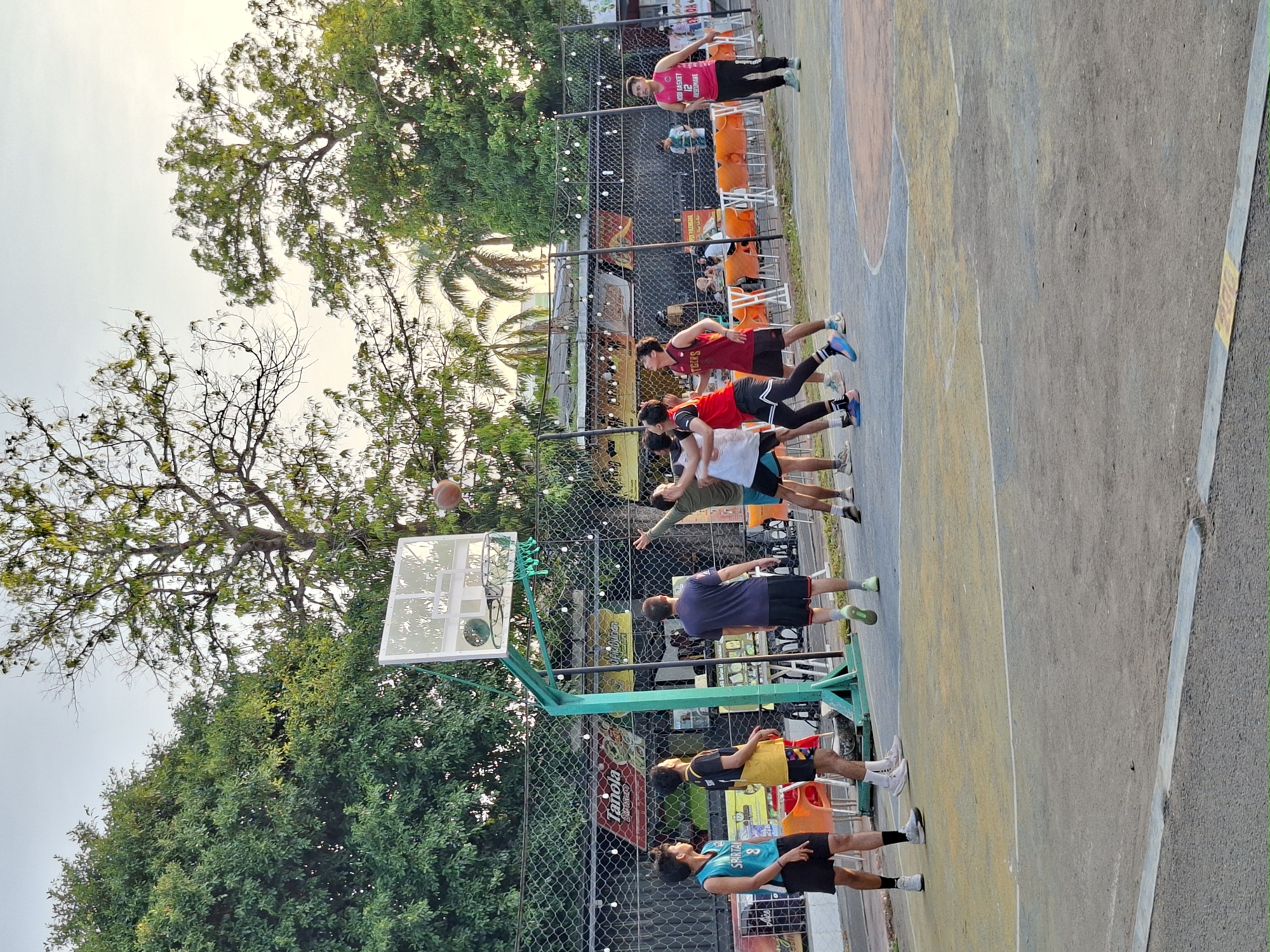 The image depicts a group of men playing basketball on an outdoor court. The court is located adjacent to a fence, behind which are trees, a restaurant, and additional outdoor seating. In the foreground, a group of men stand in a line facing the basketball hoop; one man is in the process of shooting the ball. Other men on the court are observing. The location is assumed to be in the city of Lhokseumawe, Indonesia.