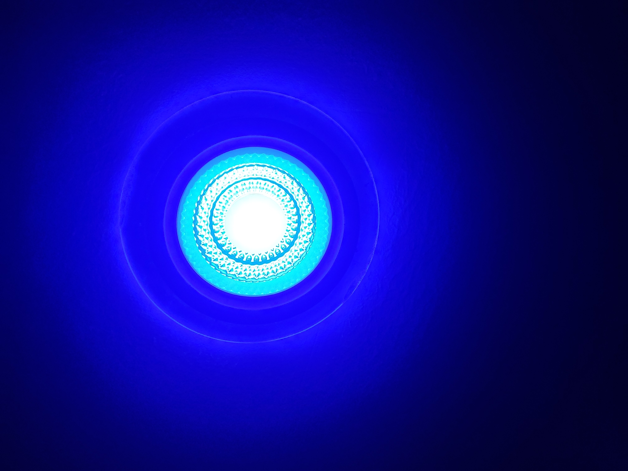 A close-up, overhead, eye-level shot captures the illuminated interior of a light fixture against a dark blue background. The light fixture itself is circular, with concentric rings of light radiating outwards. The innermost ring is a bright white, surrounded by rings of turquoise, light blue, and finally, dark blue. The texture of the light source is visible in the innermost rings, appearing to be a series of small, intricate patterns. The light is diffused, creating a soft glow that extends outwards, diminishing in intensity as it fades into the dark blue surroundings. There are no people or other objects visible in the frame. The environment is dark and nondescript.