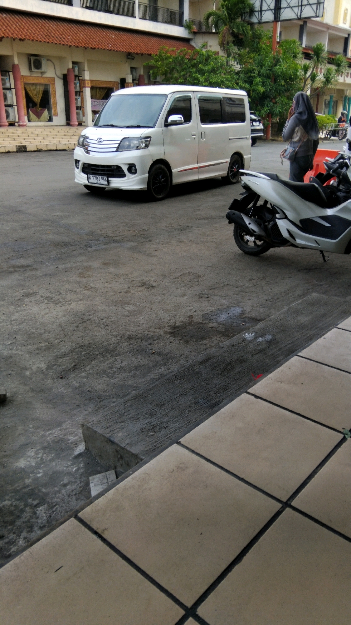 A white van is parked in an outdoor area with a concrete ground. A person wearing a hijab is standing near the van, talking on a mobile phone. In the foreground, a white scooter is parked. The background shows a building with a tiled roof and trees. The license plate of the van reads "B 2783 PKK". The scene appears to be in Pekalongan, Indonesia, based on the license plate format which is common in Indonesia.