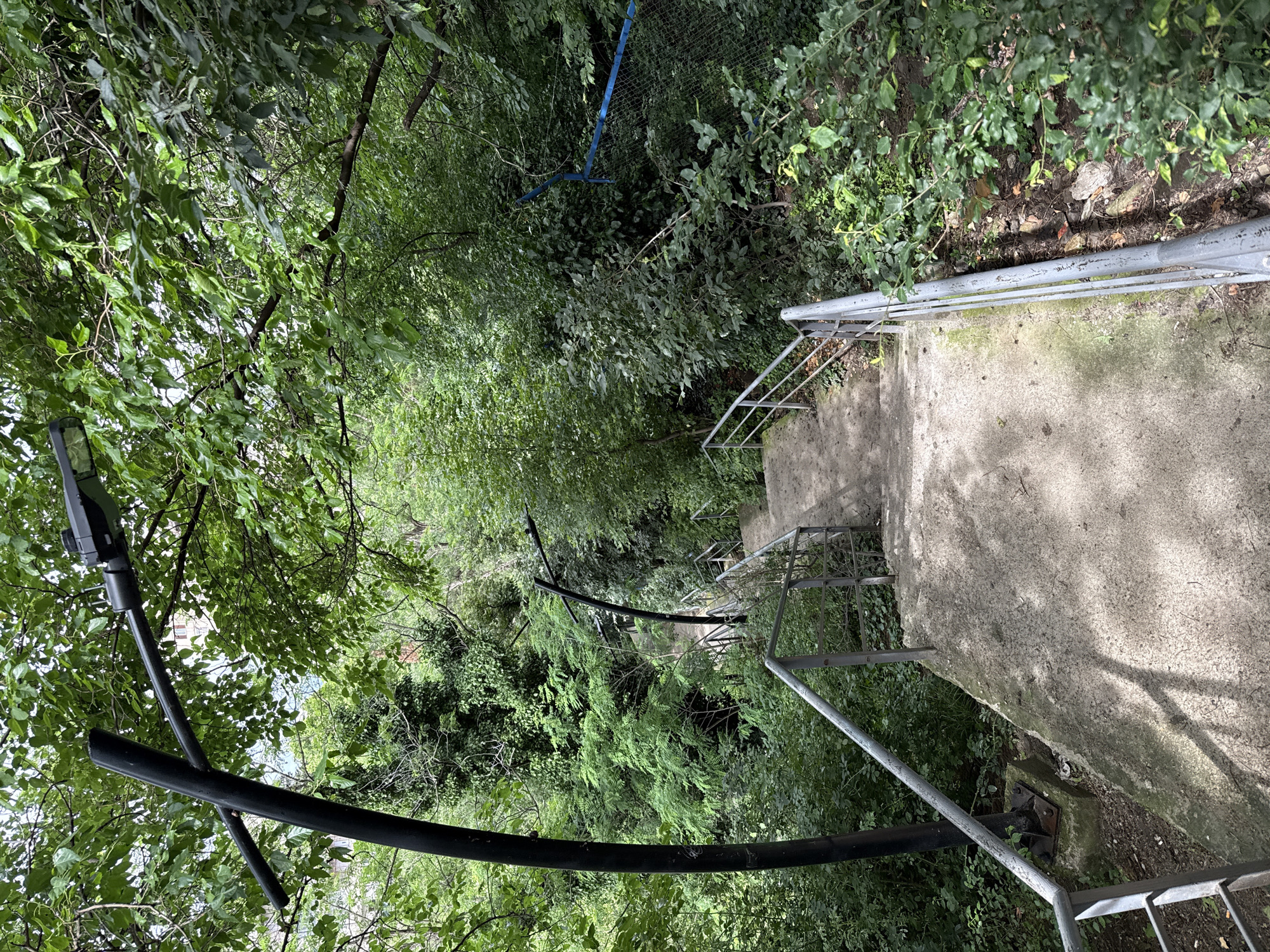 The image shows a concrete staircase descending through a dense, green, wooded area. The staircase has metal railings on both sides. A black lamppost is visible in the upper left quadrant of the image, and another, curved lamppost is seen further down the stairs. Trees and foliage heavily surround the pathway, with some leaves creating dappled shadows on the stairs. A blue fence can be seen in the background to the right. The scene appears to be an outdoor public space, possibly a park or a pathway in a residential area. There are no people visible in the image.