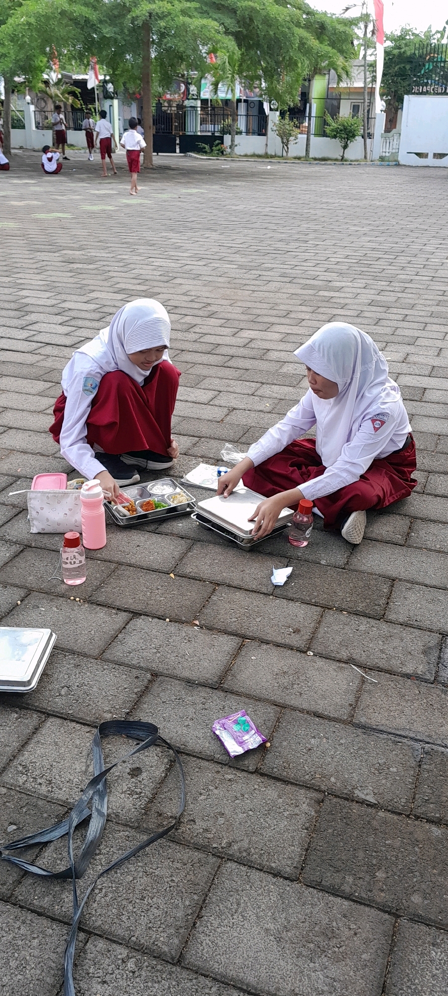 Two young girls in white headscarves and maroon skirts are sitting on a paved outdoor area, likely a schoolyard in Rogojampi, Indonesia. One girl is looking down at a metal food tray with compartments containing various food items and is holding a white paper. The other girl is looking at a stack of metal trays. In front of them are a pink water bottle, a pink lunchbox with a pattern, and a clear plastic bottle. The ground is paved with rectangular stones, and there are some scattered items including a purple packet, a black plastic strap, and some small white pellets. In the background, other children in similar school uniforms are visible, some standing, some sitting, and one appears to be walking away. Trees with green leaves and a building are also in the background, with Indonesian flags visible.