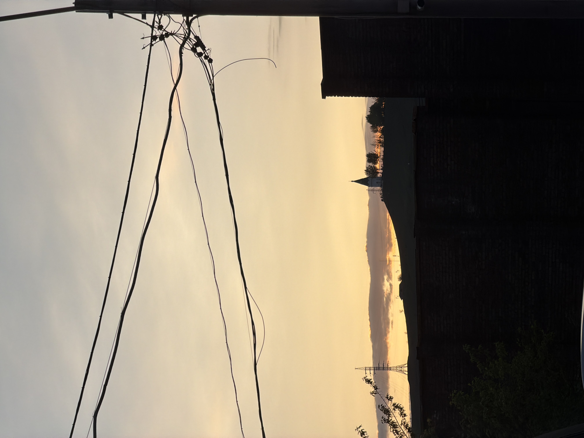 The image depicts an urban sunset scene in Krasnoyarsk, Russia, characterized by a strong contrast between light and shadow. Power lines dominate the left foreground, their intricate web of wires silhouetted against the fading sky. A dark brick building occupies the right side of the frame, its form only partially visible. In the distance, a church spire and a row of trees are faintly illuminated by the setting sun. An electrical transmission tower stands near the horizon, a testament to the urban infrastructure. The overall scene captures the intersection of nature and human construction in a cityscape.