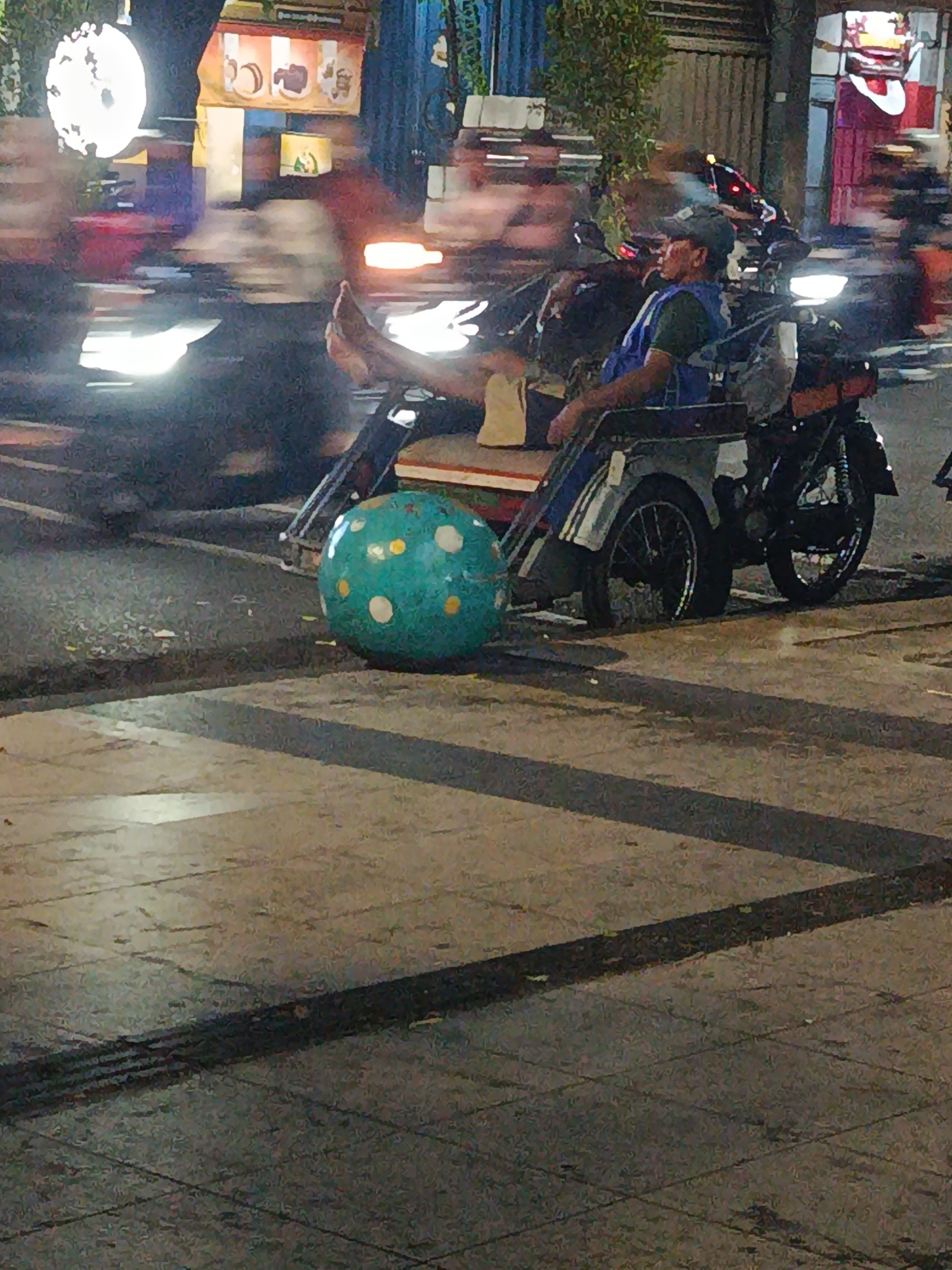 A low-angle, long shot captured at night on a sidewalk in Surabaya, Indonesia, shows a man sitting in a motorized rickshaw. The man, wearing a dark green t-shirt and a blue vest over it, has a dark baseball cap on and his legs are extended. Beside the rickshaw is a large, turquoise ball with white and yellow polka dots. In the background, traffic is visible, with blurred headlights and taillights indicating movement. The scene suggests an urban environment with pedestrian and vehicular traffic.