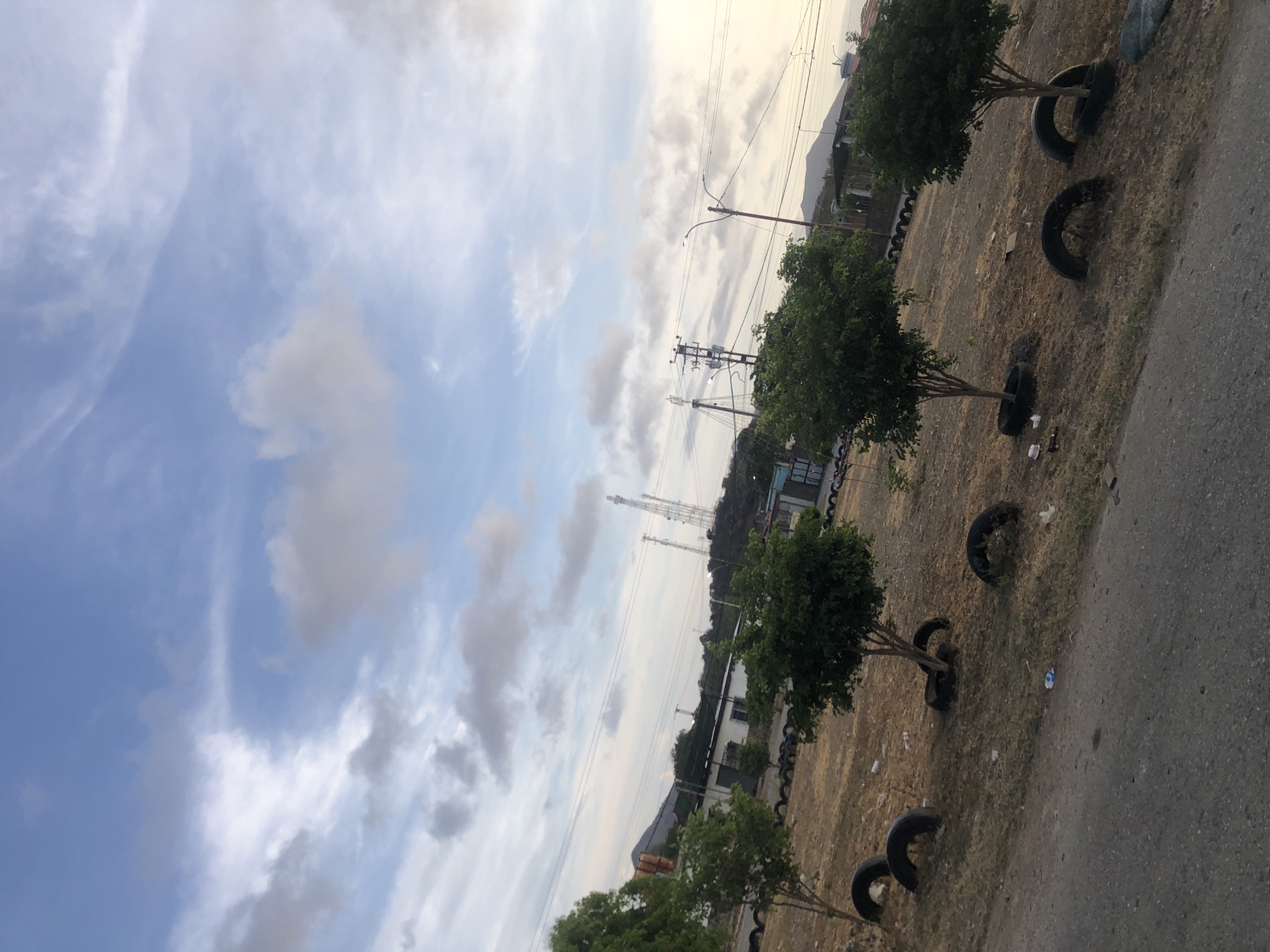 The image shows a street scene in Altagracia, Venezuela. The foreground features a line of small trees, each surrounded by several tires. Behind the trees is a dry, grassy area. A street runs along the right side of the image. In the background are buildings, power lines, and communication towers. The sky is cloudy.