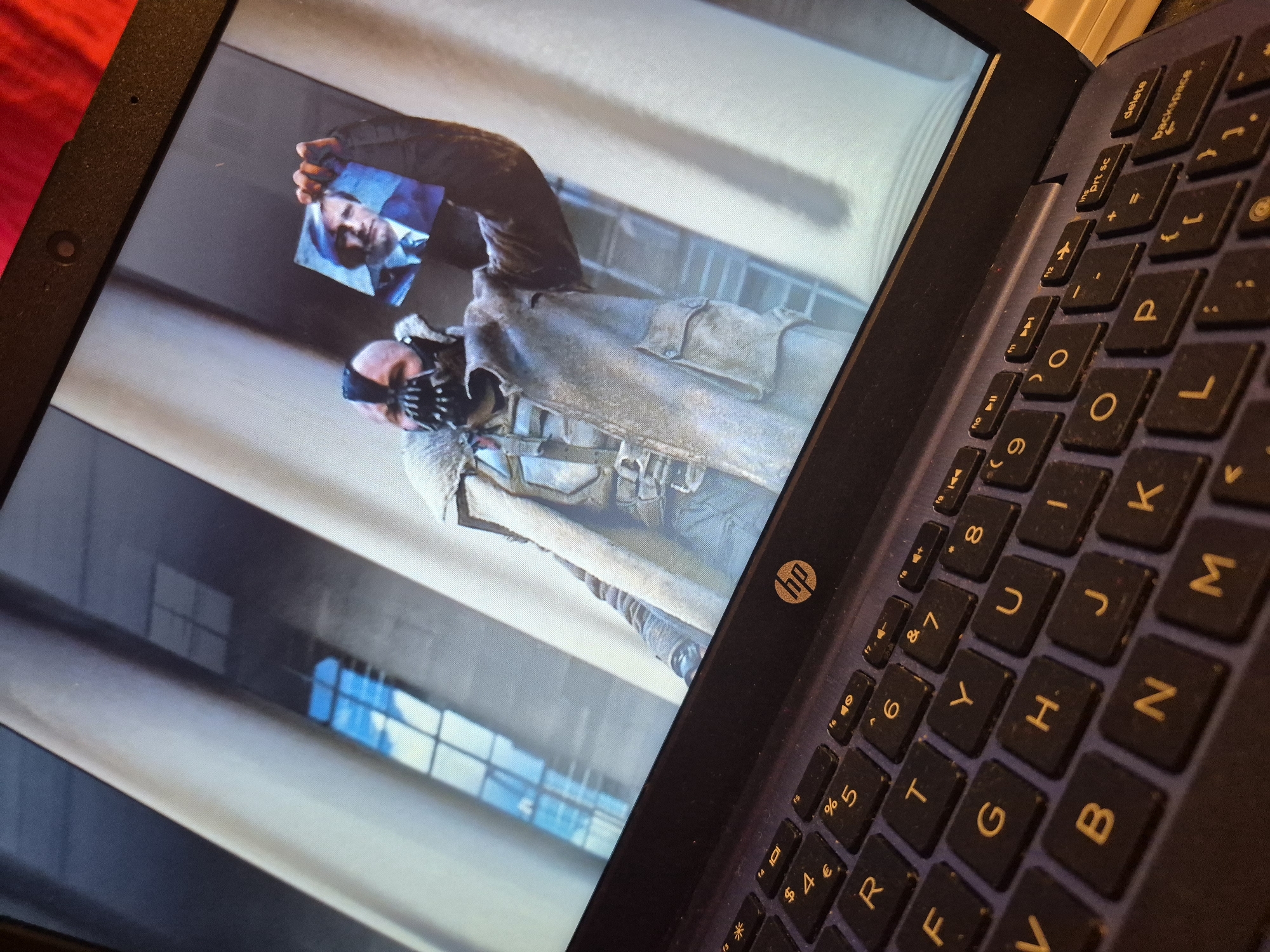 A close-up, low-angle shot shows a laptop screen displaying a scene from the movie "The Dark Knight Rises." A character, identified as Bane, is holding a small photograph of a man in a suit. The laptop's keyboard is visible in the foreground, with the HP logo present on the laptop. The background of the movie scene features large, classical-style columns and a window with a grid pattern. The overall setting appears to be indoors, likely a film set. The image's metadata or any visual cues do not indicate a specific location within the city of Sandwell, United Kingdom.