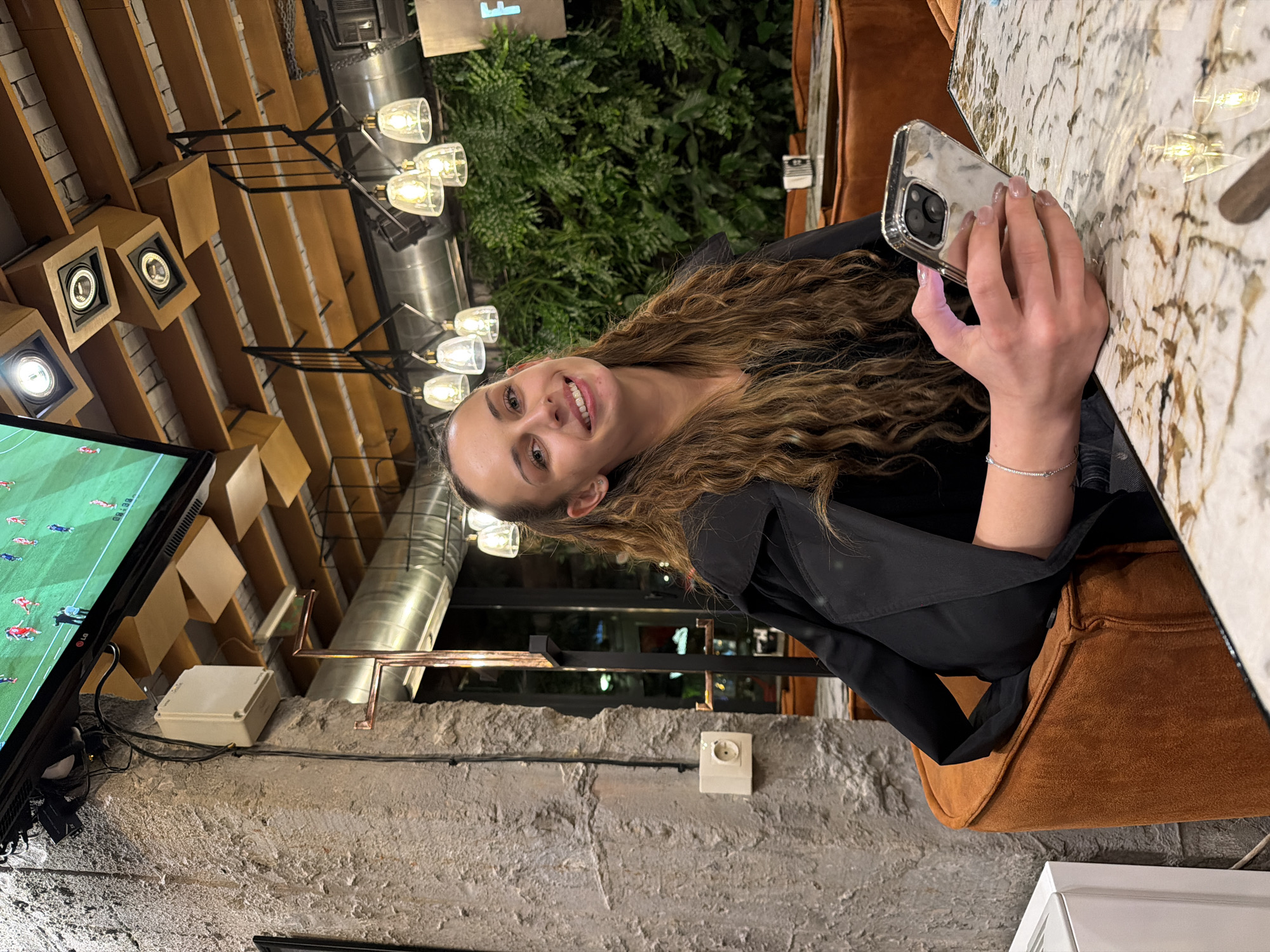 A young woman with long, wavy brown hair is captured in an indoor setting, likely a cafe or bar in Sofia, Bulgaria. She is smiling cheerfully at the camera, conveying a relaxed and happy mood. Her right hand, adorned with a delicate silver bracelet on her wrist, holds a smartphone with a reflective, possibly metallic, case. She is seated at a marble-patterned tabletop.
The establishment features a distinctive interior design combining industrial and natural elements. In the background, a large flat-screen TV displays a soccer match, showing players on the field and what appears to be a score or timer "08 30." The ceiling above the TV is notable for its wooden slat design, featuring integrated speakers within wooden boxes. The space is illuminated by stylish multi-bulb chandeliers with clear glass shades, creating a warm, ambient light, suggesting an evening or nighttime setting.
Behind the woman, a vibrant wall of green foliage adds a touch of nature to the urban environment. The walls are made of exposed, rough-textured concrete, complemented by visible metal pipes and conduits, enhancing the industrial aesthetic. An electrical outlet and a small white junction box are also visible on the wall. The woman is dressed in a black top or jacket and is seated in a comfortable brown upholstered chair or booth.