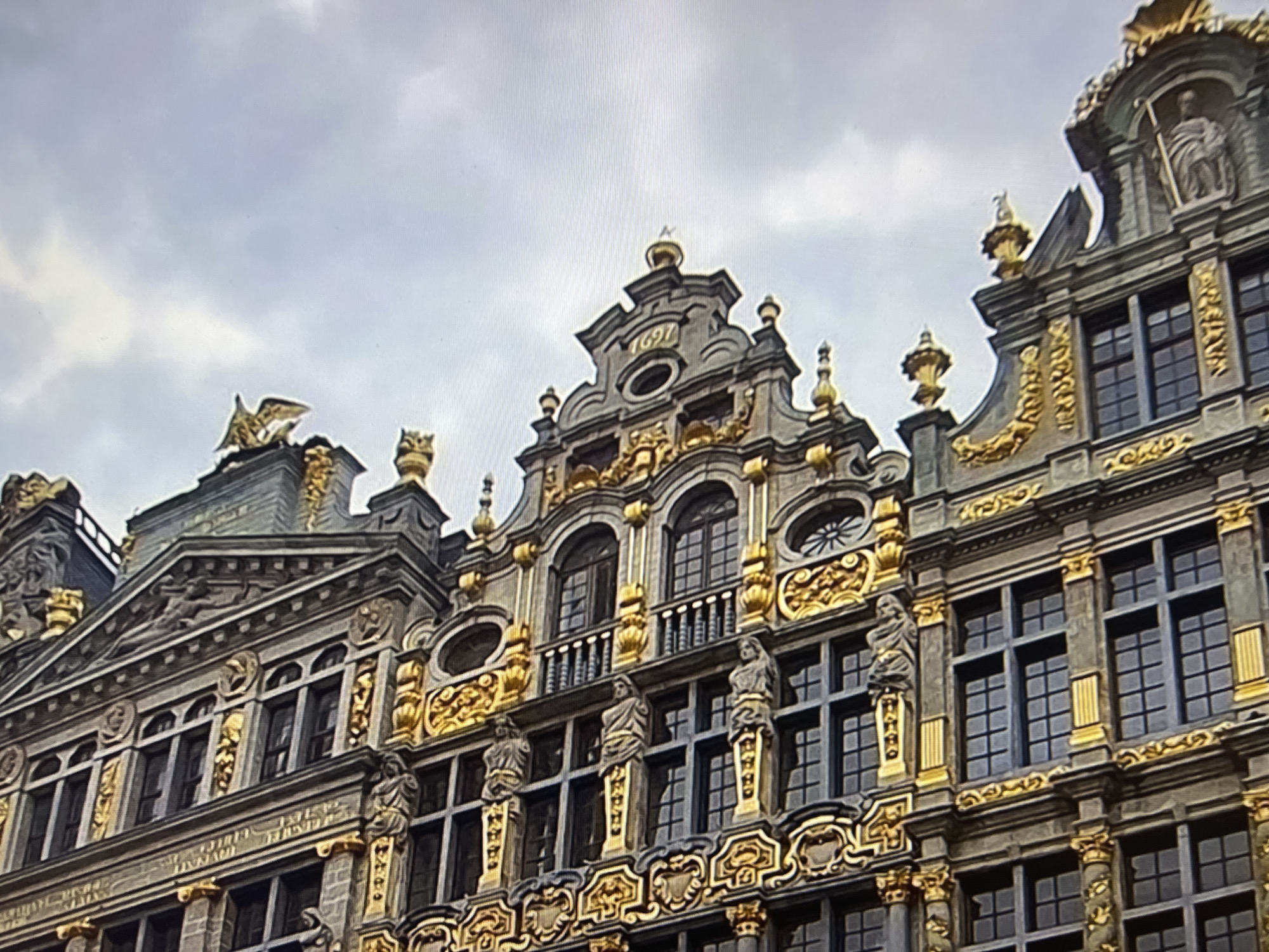 The image shows a detailed view of the facade of an ornate historical building, characterized by Baroque or Rococo architectural style. The building features a grey stone exterior adorned with extensive gold decorative elements. These gold embellishments include intricate carvings, moldings, statues, and gilded architectural details such as columns, cornices, and finials.
Prominent features include multiple stories with numerous windows, some of which are arched and others rectangular. Many windows have decorative golden frames and grilles. On the upper levels, sculptures of figures are visible, some standing and others integrated into the architectural design. The roofline is complex, with pediments, gables, and decorative spires. One of the gables prominently displays the year "1697" in gold numerals. A golden eagle sculpture is perched atop a pinnacle on the left side of the building. The sky is overcast with grey clouds. The image is a close-up, focusing on the architectural details of the building.
There is no visible connection to the city of Qarağandı city administration, Kazakhstan, in this image. The architectural style and details are not characteristic of that region, and there are no indicators in the image that would place it there.