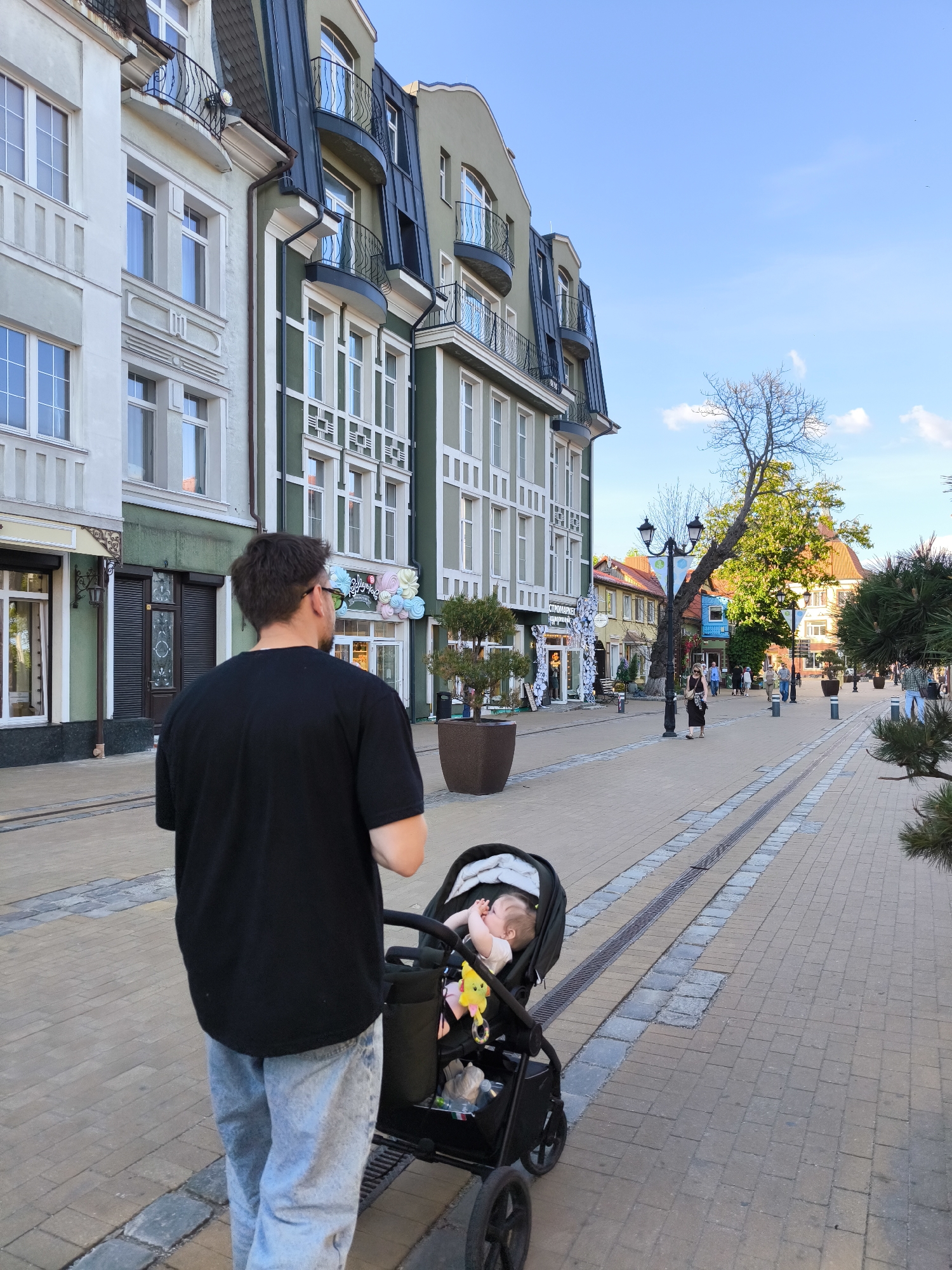 A man is seen from behind, walking along a sunny pedestrian street, pushing a baby in a stroller. He is wearing a black t-shirt and light blue jeans. The baby, partially visible in the stroller, appears to be sleeping or resting with its hands near its face.
The street is paved with light-colored cobblestones, featuring two central lines of darker grey stones, possibly remnants of old tracks or decorative elements. Tall, ornate streetlights are spaced along the street. On both sides, multi-story buildings line the street. The buildings on the left have light-colored facades (white, cream, and light green) with decorative window frames and dark, sloped roofs featuring dormer windows and curved balconies with dark railings.
In the background, other people are strolling along the street, and there are various shops and businesses. A large tree with bare branches and some green foliage stands further down the street on the right. The sky is clear and blue with a few scattered white clouds, indicating a bright daytime, likely a pleasant afternoon.
Visible text on a shop sign on the left reads "СТРОЙМАРКЕТ" (Stroymarket). Further down, another shop sign displays "Sillypops" next to colorful decorations. The architecture and street design suggest a European town setting, consistent with Zelenogradsk, Russia. The overall atmosphere appears peaceful and leisurely.