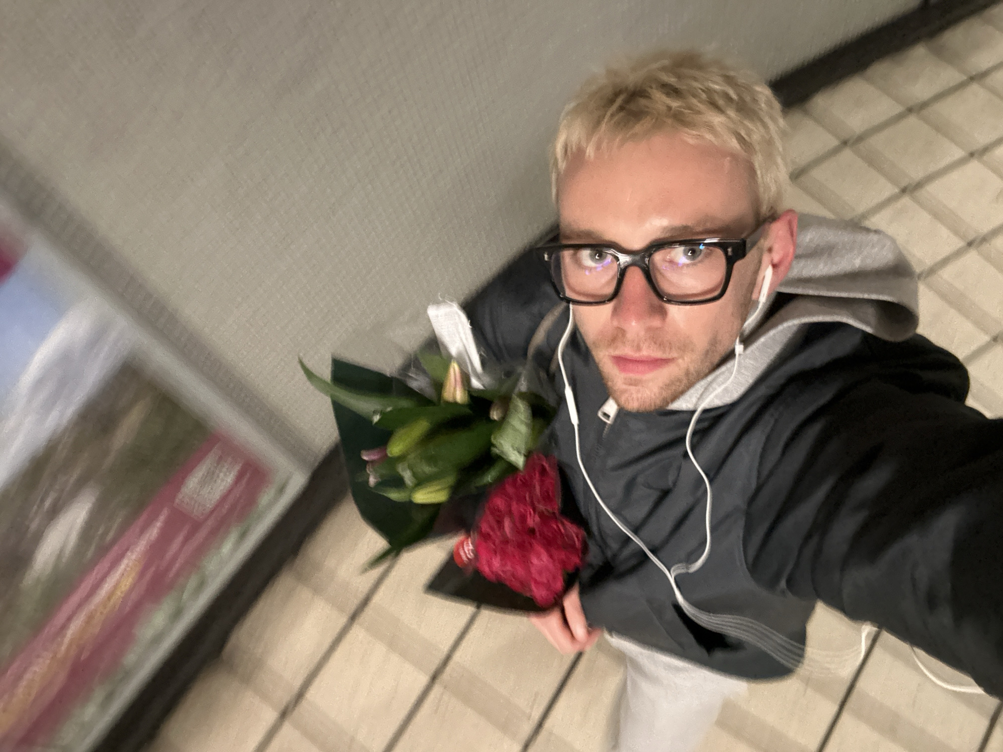 A person with blonde hair and glasses is holding a bouquet of flowers in a selfie. The person is wearing a dark jacket over a gray hooded sweatshirt and white pants. The flowers consist of red carnations and what appears to be lilies. The person has earphones in their ears. The background includes a beige tiled floor, a beige wall and a blurred display or window to the left. The location is not recognizable as being in the city of London, United Kingdom.