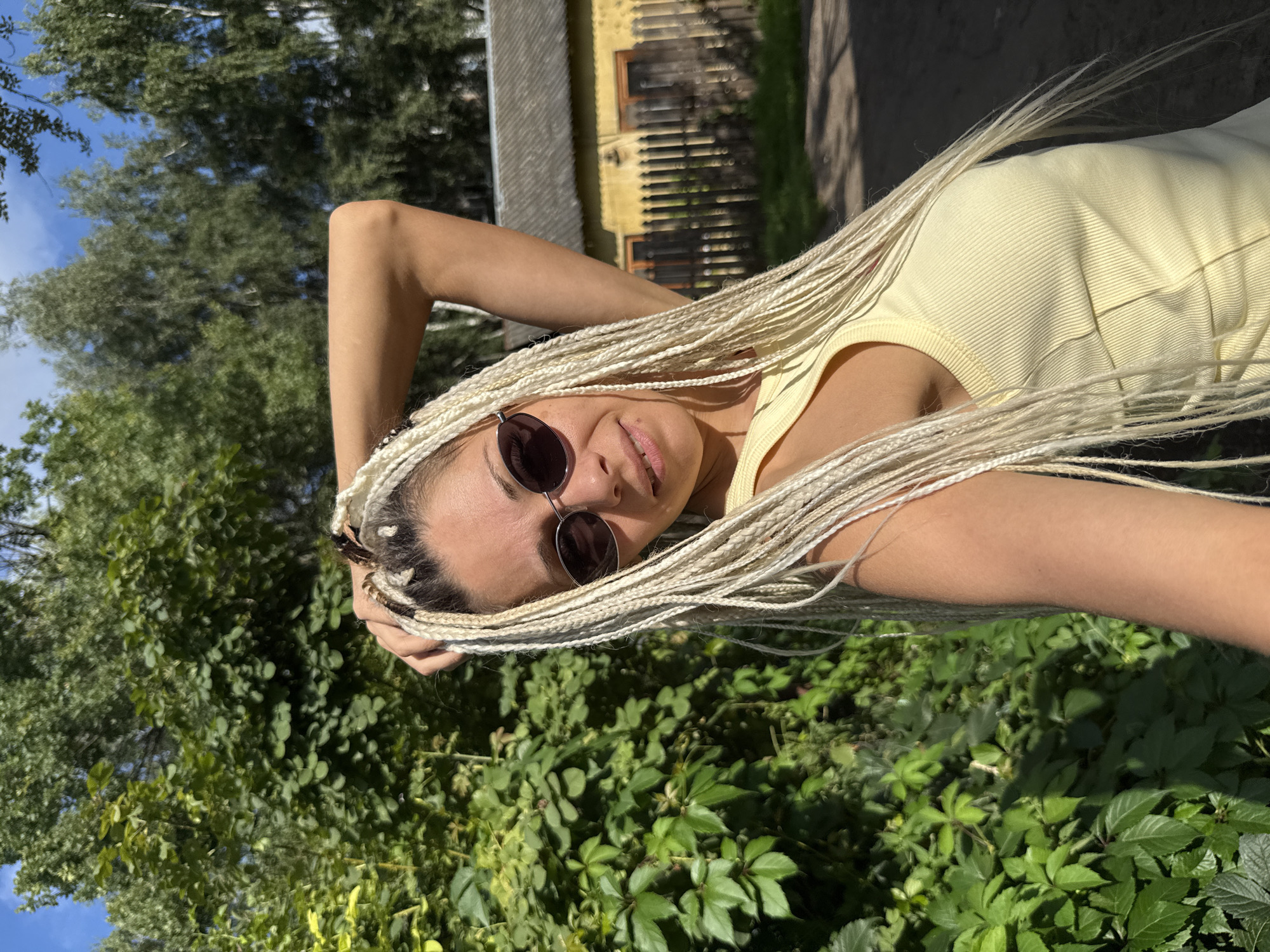 A woman with blonde braided hair and sunglasses poses for a selfie outdoors, with lush green foliage on her left and a weathered yellow building with a wooden fence in the background. The scene appears to be set in a rural or suburban area of Rusanovo, Russia, indicated by the architecture and the natural surroundings. The woman is wearing a yellow tank top and has her right arm raised, touching her hair. The lighting suggests it is a sunny day, with shadows cast on the ground.