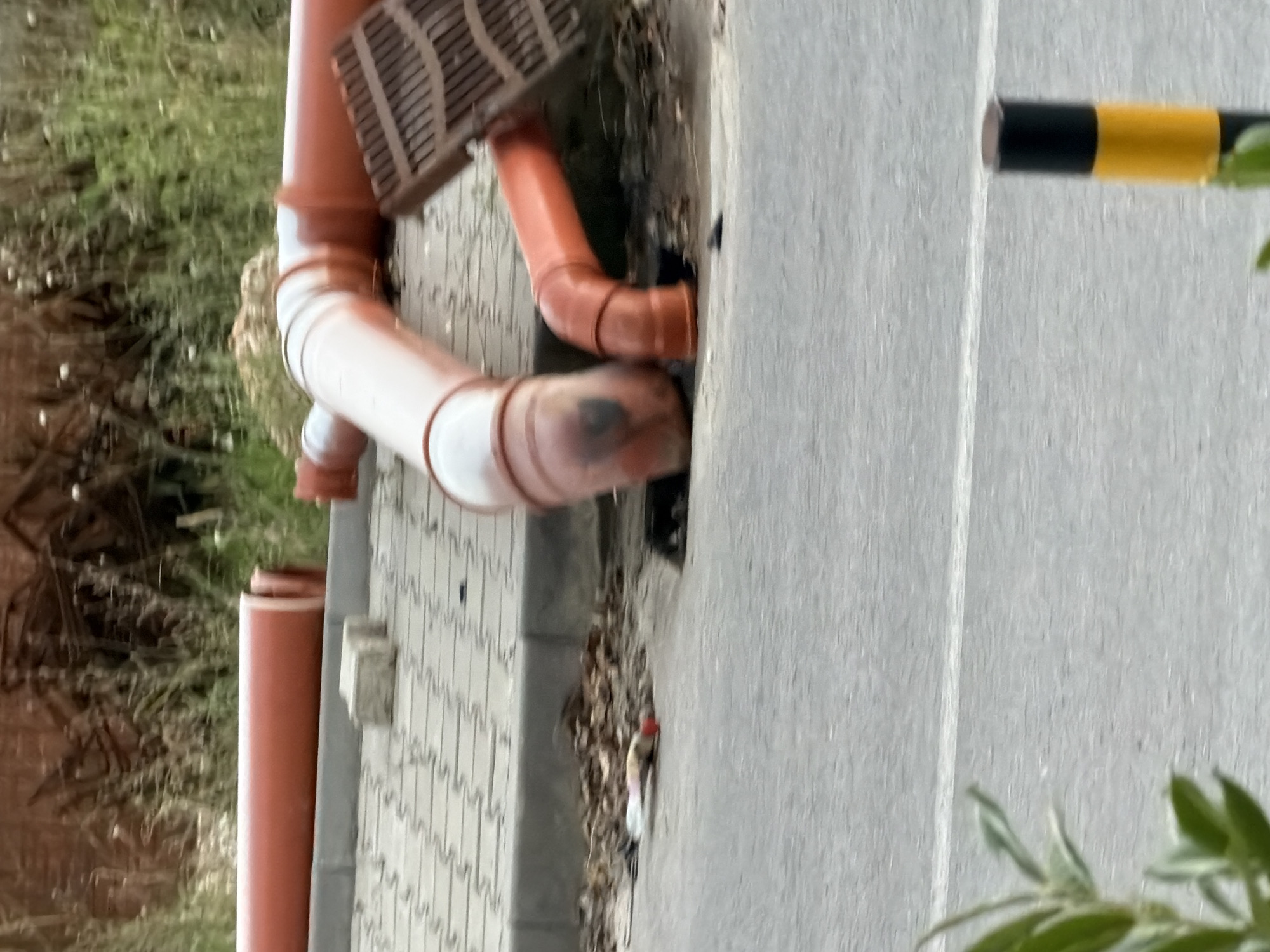 The image depicts a construction site with orange pipes laid out on a paved area next to a road. The pipes are of varying sizes and appear to be part of a drainage or utility system. One pipe is connected to a metal grate. In the background, there is overgrown vegetation and some debris. In the foreground, a traffic bollard with yellow and black markings is visible on the side of the road. The scene is set in a somewhat urban environment, potentially in Plovdiv, Bulgaria, given the context. There are no people visible in the image, and the focus is on the construction materials and the surrounding environment. The lighting suggests it is daytime.