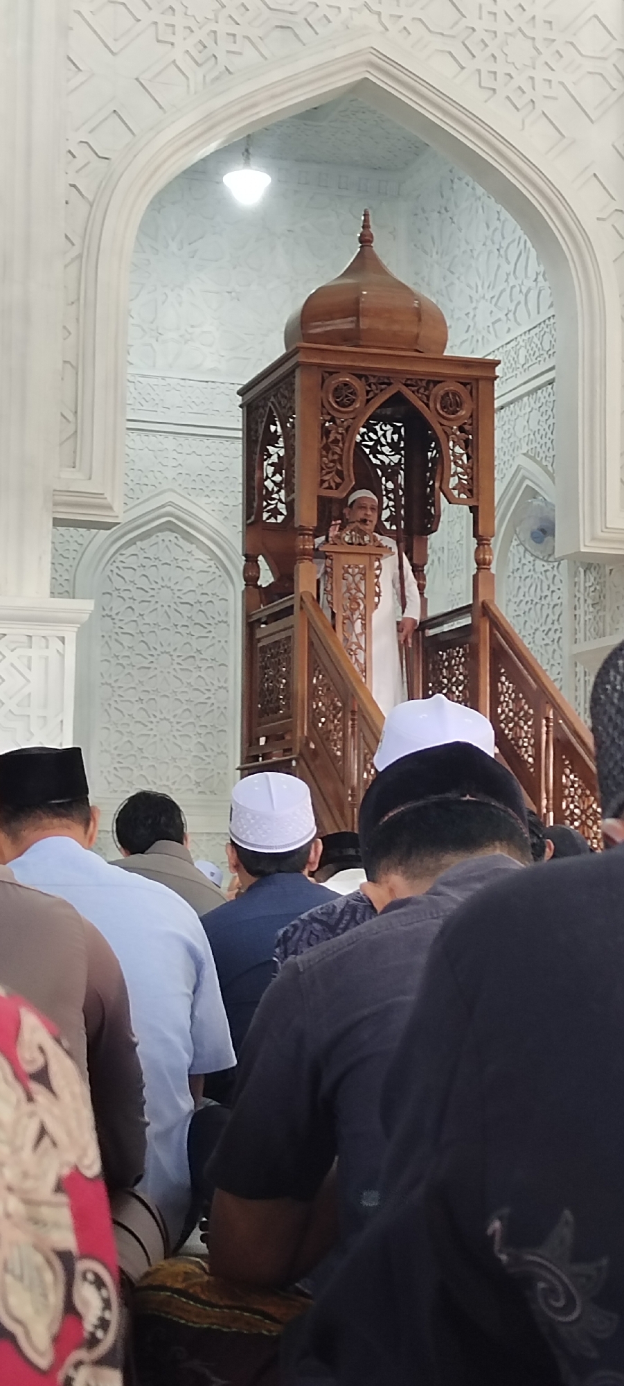 A man in white stands in a wooden pulpit, giving a speech to a group of men in a mosque. The men are wearing various colors and traditional hats, while the pulpit is dark wood with ornate carvings. The mosque's interior is predominantly white with geometric designs, including an arched doorway. The scene is likely taking place in Lhokseumawe, Indonesia.