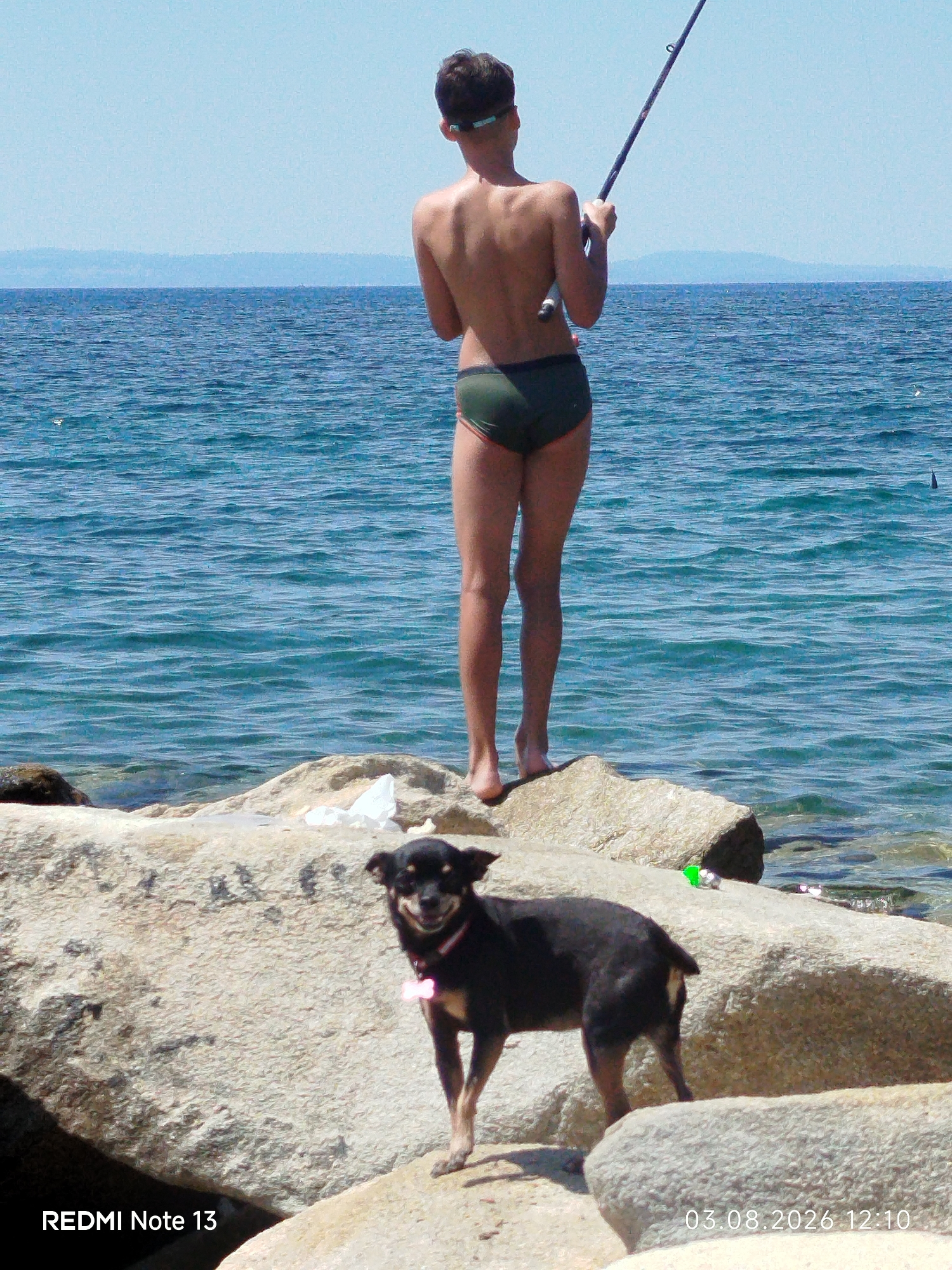 A boy, facing away from the camera, stands on a rocky shore near Sithonia Municipal Unit, Greece, fishing in the turquoise sea. He is wearing dark green swim trunks and goggles pushed up on his forehead. A small black dog with a pink collar stands on a large rock in the foreground, looking towards the camera with its tongue out. The sea is calm with gentle waves, and the sky is clear and blue. The rocky shore is made up of large, light-colored boulders. The image has a timestamp in the bottom right corner: 03.08.2026 12:10.