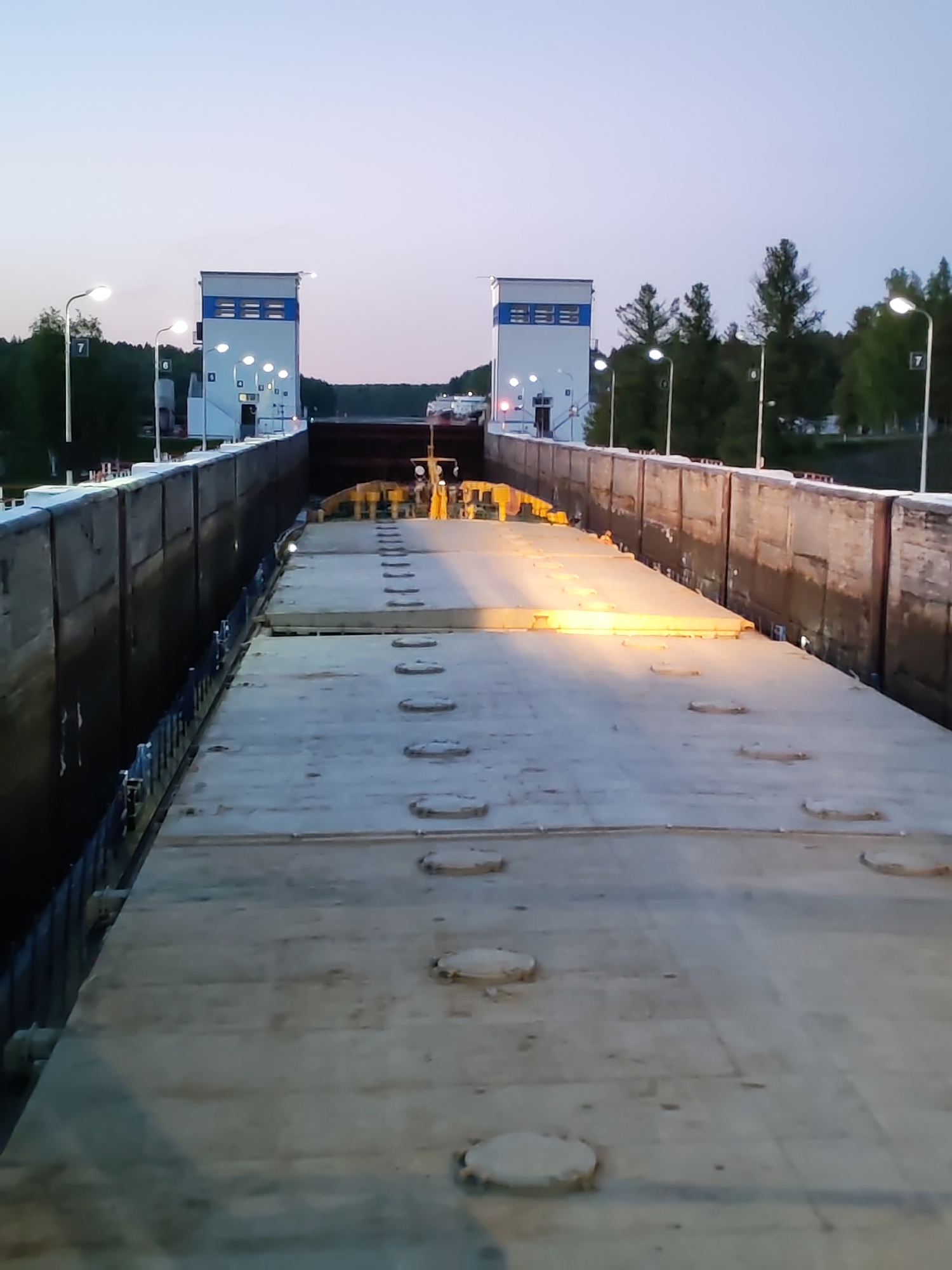 This image depicts a wide, flat concrete surface, likely the deck of a large barge or a section of a canal lock, viewed from inside the lock chamber at what appears to be dusk or dawn. The surface extends into the distance and is marked with numerous circular, raised covers, resembling manholes or inspection ports. A warm yellow light illuminates a portion of this concrete in the mid-ground.
Tall, dark concrete walls flank the central area on both the left and right, forming the sides of the lock. Some blue-colored railings or fittings are visible along the base of these walls.
In the background, two identical, multi-storied control towers or buildings stand at the far end of the lock. These structures are white with prominent blue horizontal stripes and are brightly illuminated by multiple lights. Behind and around the towers, dense green trees indicate a natural or semi-rural setting. The sky above is a soft gradient of light blue and pinkish-purple, typical of twilight.
Artificial streetlights, featuring bright, circular luminaires, are visible along the tops of the lock walls and near the control towers, providing additional illumination to the scene. On a lamppost on the right side, the number "7" is clearly visible, and another lamppost on the left appears to show a single digit, possibly "0" or "6". No people are visible in the frame. The scene suggests a calm, operational infrastructure, possibly in the process of a vessel transiting the lock.