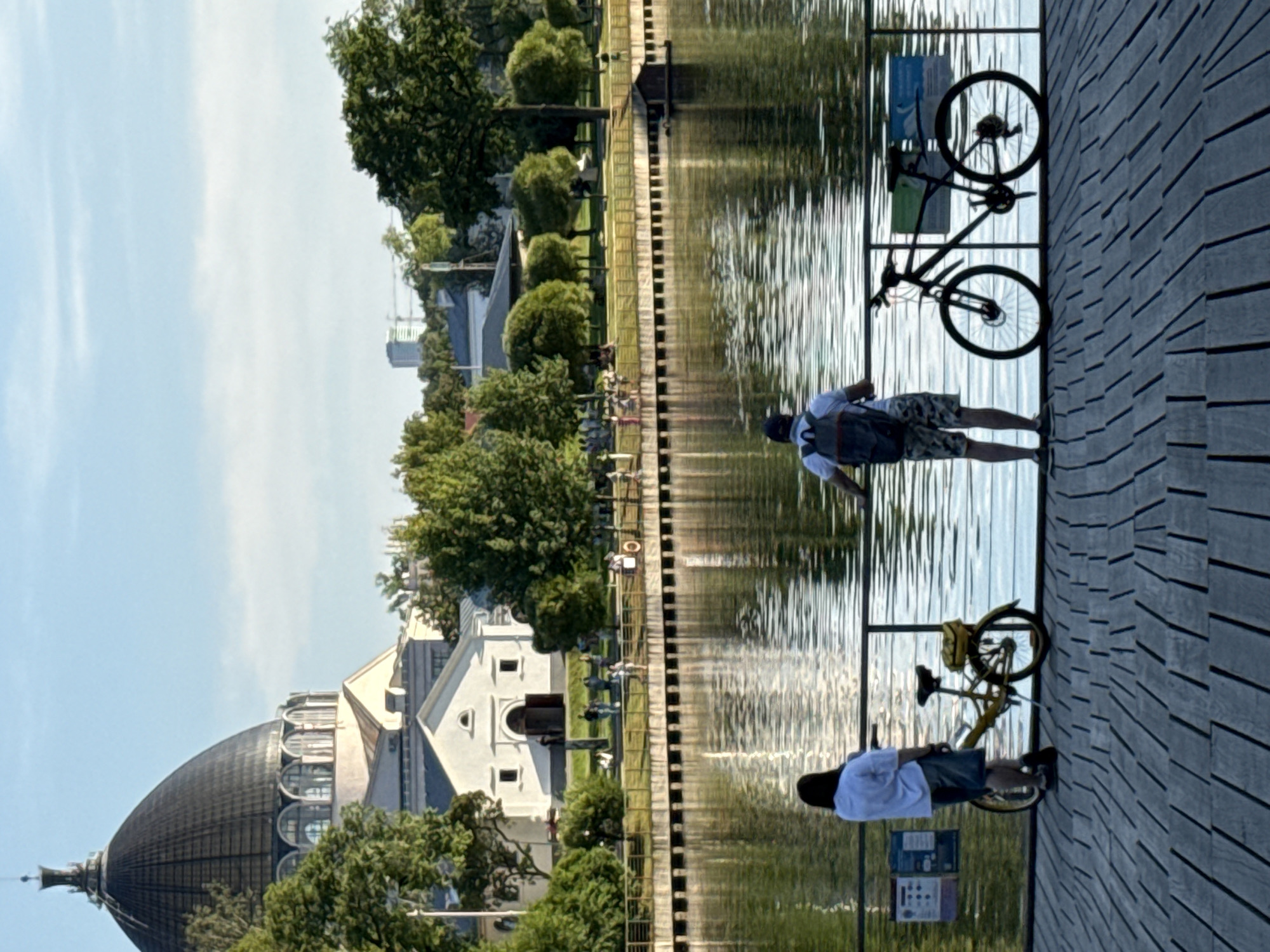 This image captures a serene view of the Moscow Planetarium's dome, a landmark in Moscow, Russia, as seen across a calm body of water. In the foreground, two individuals stand on a wooden boardwalk, each with a bicycle. The person on the left, viewed from the back, wears a light blue shirt and dark shorts, with a yellow bicycle parked beside them. The person on the right, also viewed from the back, has a backpack and is wearing camouflage shorts, with a black bicycle next to them. The boardwalk overlooks the water, which reflects the clear blue sky and the surrounding greenery. Further in the background, across the water, is a white building with a prominent arched entrance, and beyond that, more trees and structures, including the distinctive dome of the planetarium with its circular windows. The scene is bathed in the warm light of what appears to be late afternoon or early evening.