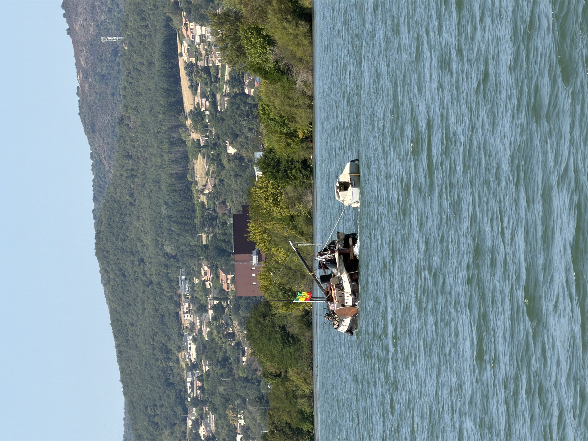 In Saint-Georges-les-Bains, France, on a body of water, two boats are visible. The larger boat, a barge-like vessel with a weathered hull, is partially submerged and appears to be listing. A flag with yellow, green, and red stripes is attached to a mast on the barge. A smaller, white boat is tethered to the larger vessel and floats independently. The background shows a densely wooded hill dotted with houses, indicating a suburban or rural setting. The sky is clear and blue. The water is a calm blue-green, with small ripples.