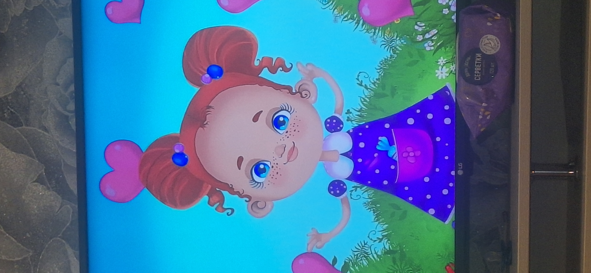 The image depicts a cartoon drawing of a young girl with red hair styled in two buns, adorned with blue beads. She has large blue eyes, freckles on her nose and cheeks, and is wearing a white collar over a purple polka-dot dress. A purple pocket on her dress contains a small blue bird figurine. She is standing in a green grassy area with small white flowers, against a bright blue background populated with pink heart shapes. Below the drawing, on a shelf, is a package of wet wipes with Russian text. The shelf is in front of a television screen displaying the cartoon. The image does not contain any information about the city of Bila Tserkva, Ukraine.