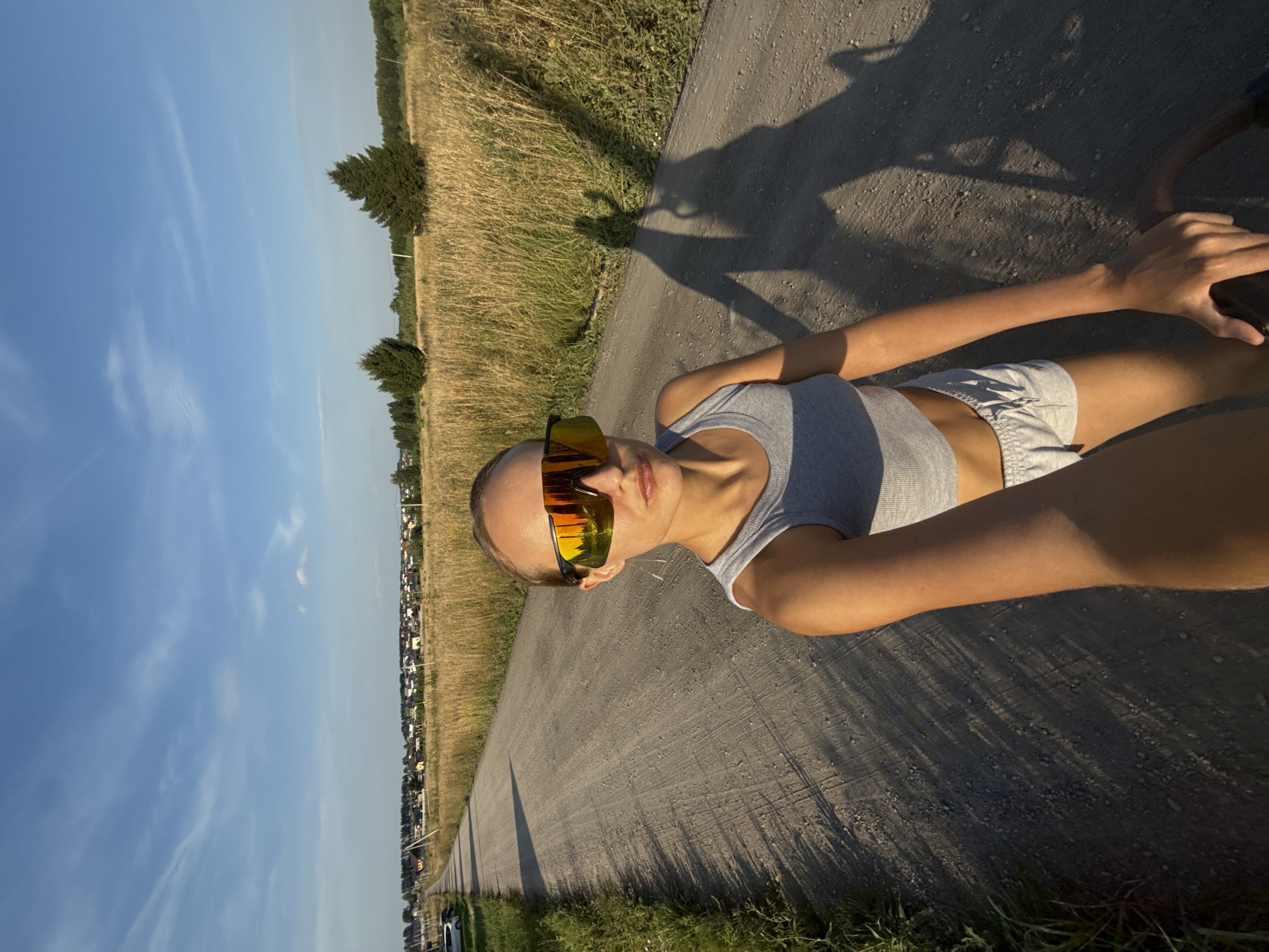 A young woman wearing a gray tank top and gray shorts, with large, reflective sunglasses, stands on a dirt road in a rural area. The road is lined with dry grass and a few scattered trees. In the distance, a small village with houses and power lines is visible. The sky is blue with a few wispy clouds. The woman's shadow, along with the shadow of what appears to be a bicycle, are cast on the road. The image was taken at sunset, as indicated by the long shadows and warm lighting. The village in the distance is identified as Podshivalovo, Russia.