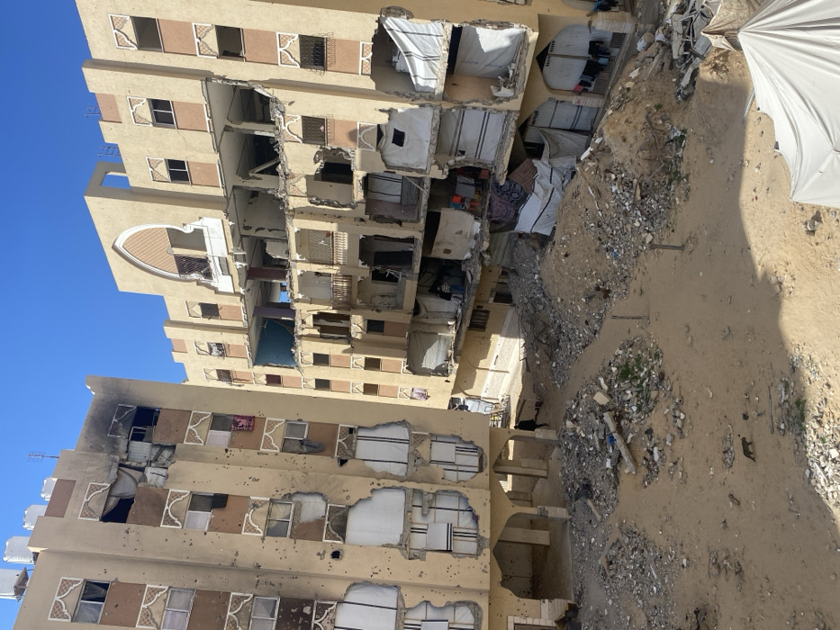 This image depicts a scene of extensive structural damage in the city of Khan Yunis, Palestinian Territory, under a clear blue sky. Two multi-story residential buildings, constructed with light beige facades and brown decorative arched elements, dominate the view. The building on the left exhibits numerous small impact holes across its surface and significant damage to its upper floors, including a blackened, burnt area on the top left, exposed interiors, and missing sections of walls. Several white water tanks are visible on its roof. Lower floors of this building also show large sections of the facade missing, revealing white panels or makeshift coverings.
The building on the right has suffered more severe, localized structural collapse, particularly impacting its middle and lower sections, which feature large, open voids where floors and exterior walls have given way. Exposed interiors reveal debris, hanging fabrics, and white tarps or sheets used as makeshift covers. Architectural arches are damaged.
The ground between and in front of the buildings is an unpaved, sandy area covered with substantial piles of concrete rubble, twisted metal, and miscellaneous debris. A dirt path traverses this debris field. One individual, dressed in dark clothing, walks on this path near the base of the left building. A small group of individuals is visible inside a damaged section of the right building on a lower floor, near a white tarp. A dark-colored cat walks on the sandy ground in the mid-ground. Makeshift white tarpaulins are visible covering damaged sections on the lower levels of the right building, and clothes are hanging to dry on a line in the lower right corner of the right building. A portion of a large white tent structure is visible in the lower right foreground. Shadows cast by the buildings and debris indicate strong sunlight.