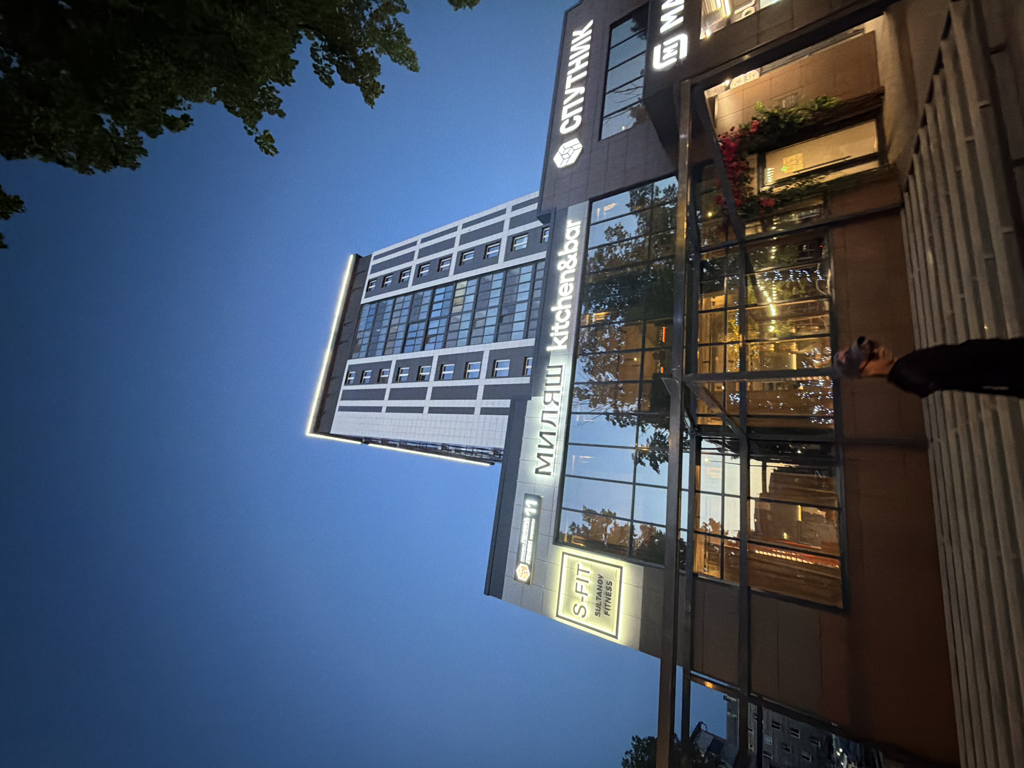 A street-level exterior shot, taken from a low angle, shows the facade of a modern multi-story building against a twilight sky. The building features a dark, sloped lower section with large glass windows reflecting the lights from within, hinting at a restaurant or bar with festive decorations. Above this is a lighter-colored, taller section of the building with a grid-like pattern of windows. A prominent illuminated sign reads "S-FIT SULTANOV FITNESS" on the lower facade, and further along, signs display "МИЛЯШ kitchen&bar" and "СПУТНИК." The scene is framed by a leafy tree on the right side. A person wearing a baseball cap and sunglasses, dressed in dark clothing, is partially visible in the foreground, walking up a set of concrete stairs. The overall impression is one of urban nightlife in Tujmazy, Russia.