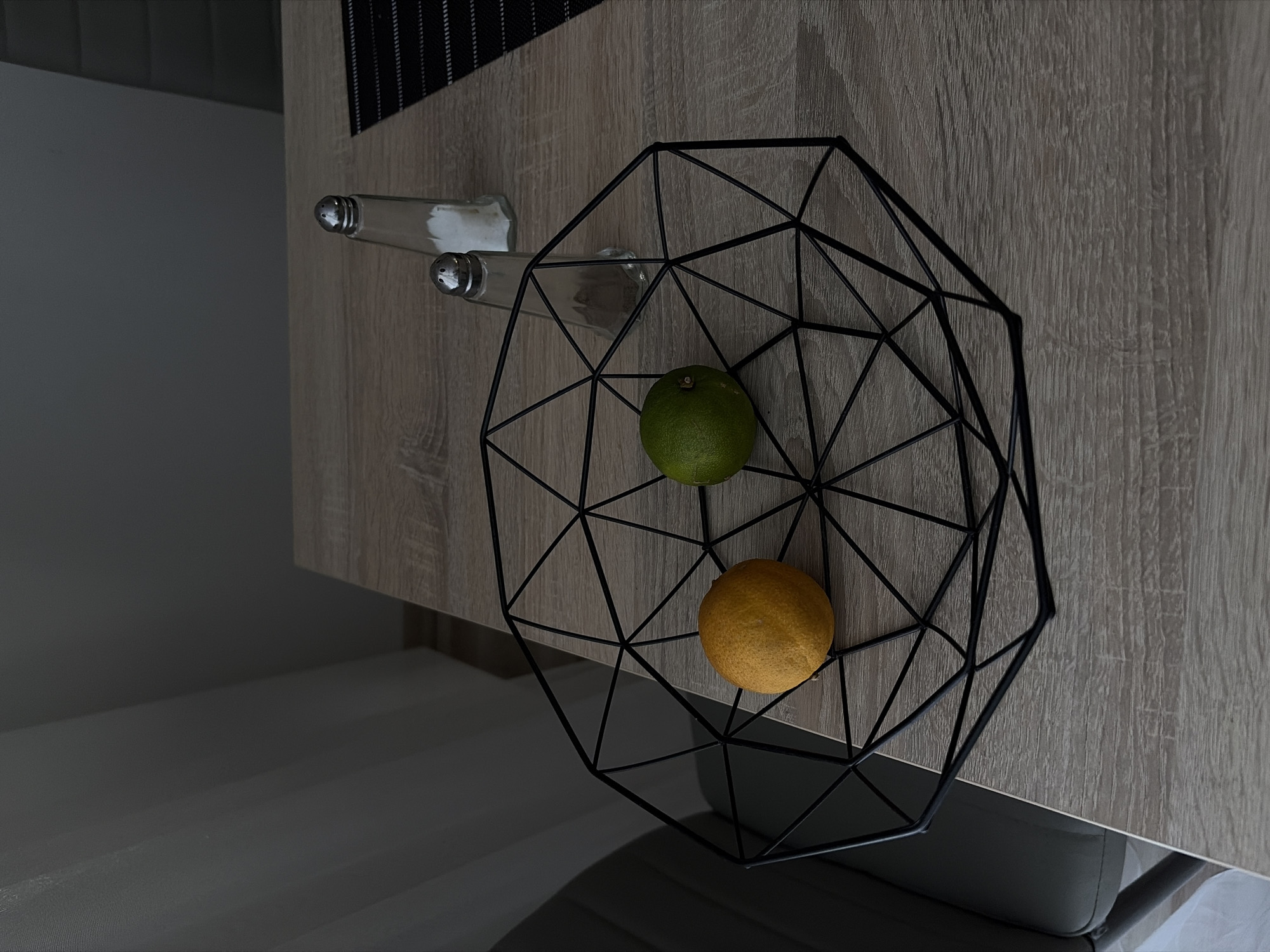 The image depicts a close-up, eye-level shot of a wooden table with a geometrically shaped black wire basket containing one yellow lemon and one green lime. Two salt and pepper shakers are positioned behind the basket. The background shows a portion of a grey chair and a sheer white curtain. The lighting suggests an indoor setting, possibly a dining area or kitchen. There are no people visible in the image. The scene is set in Nitra, Slovakia.
