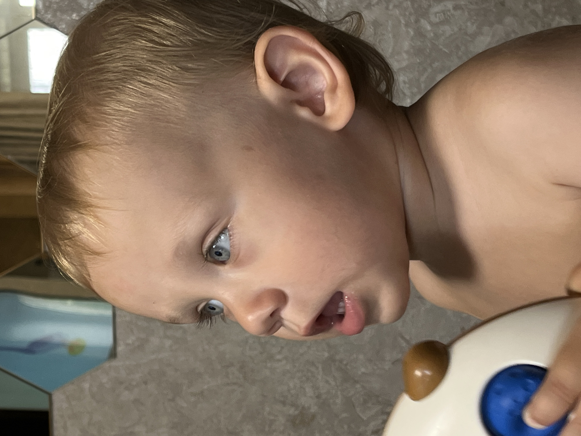 A close-up, side profile of a fair-skinned toddler with blonde hair and blue eyes is captured. The toddler's mouth is slightly open, revealing teeth, and their gaze is directed upwards and to the left. A portion of a toy, primarily white with a brown rounded protrusion and a blue circular element, is visible on the left side of the frame, being held by what appears to be a person's hand. The background consists of a textured grey wall and a reflective surface with a geometric blue and yellow design. The setting is indoors, likely a bathroom, as suggested by the tiled wall. The image does not contain any identifiable landmarks or elements that would allow for the determination of its location in Nizhny Novgorod, Russia.
