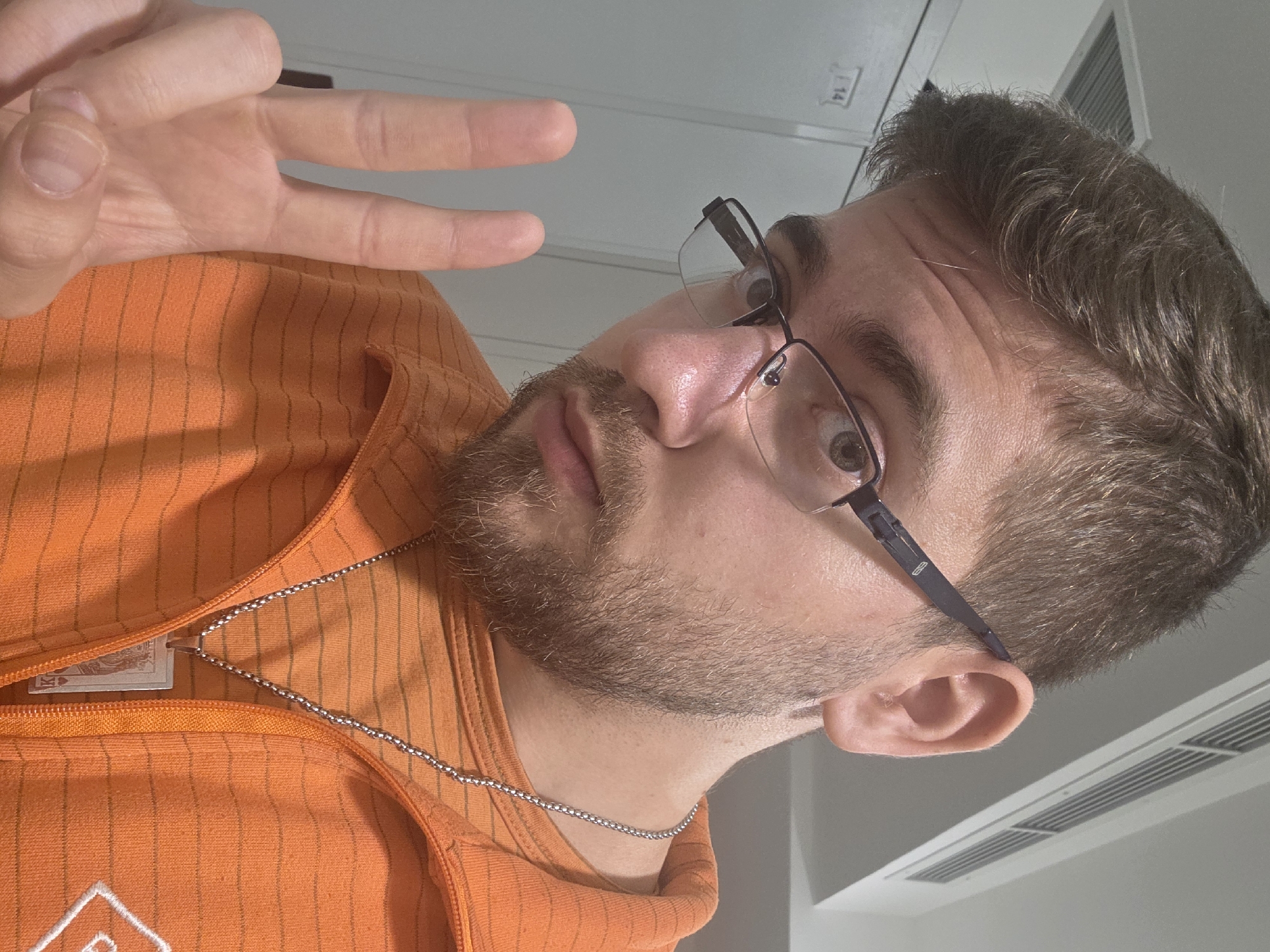 A man with brown hair and a beard, wearing glasses and an orange striped jacket, is making a peace sign with his hand. He is wearing a silver chain necklace. The setting appears to be indoors, with a ceiling fixture visible in the background. There is no information in the image that relates to the city of Plovdiv, Bulgaria.