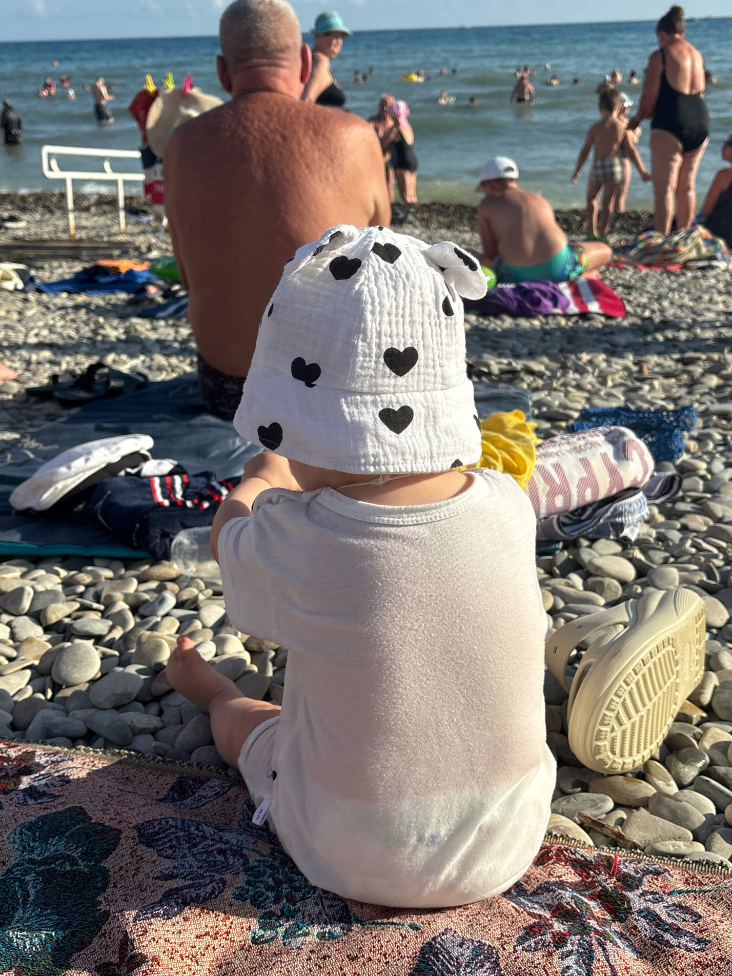 At a pebbled beach in Arkhipo-Osipovka, Russia, a toddler wearing a white sun hat with black heart patterns and a white onesie sits facing away from the camera. The toddler is positioned on a colorful blanket amidst a scatter of beach pebbles. To the right, a light-colored sandal rests on the pebbles. In the background, several adults and children are visible wading in the sea under a bright sky. Clothing items are also present on the pebbles near the toddler.