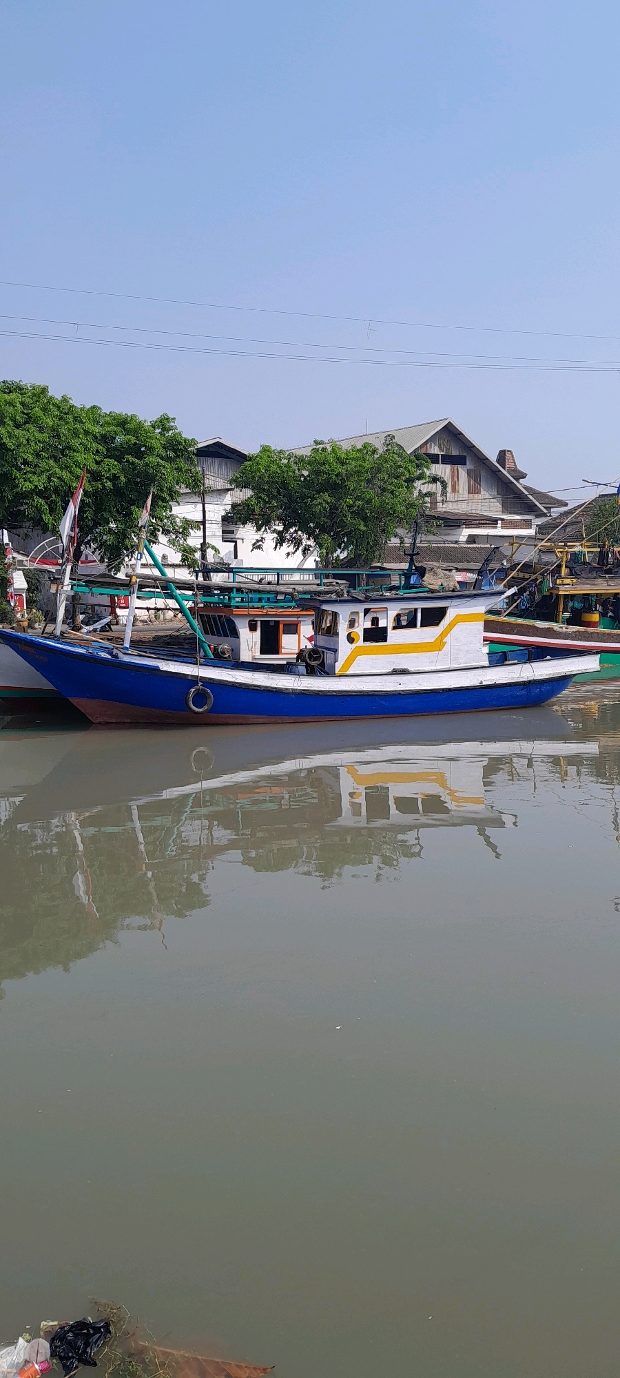In Pasuruan, Indonesia, a brightly colored fishing boat is docked in a murky waterway. The vessel is painted blue along its hull, with a white superstructure and a yellow stripe running along its side. A small cabin is visible on the deck. The boat is secured with ropes. In the background, trees with lush green foliage line the bank, behind which stand several buildings with corrugated metal roofs. The sky is clear and blue. The water reflects the boat and the surrounding environment, though the image is marred by floating debris, including plastic bags and leaves, near the bottom left corner.