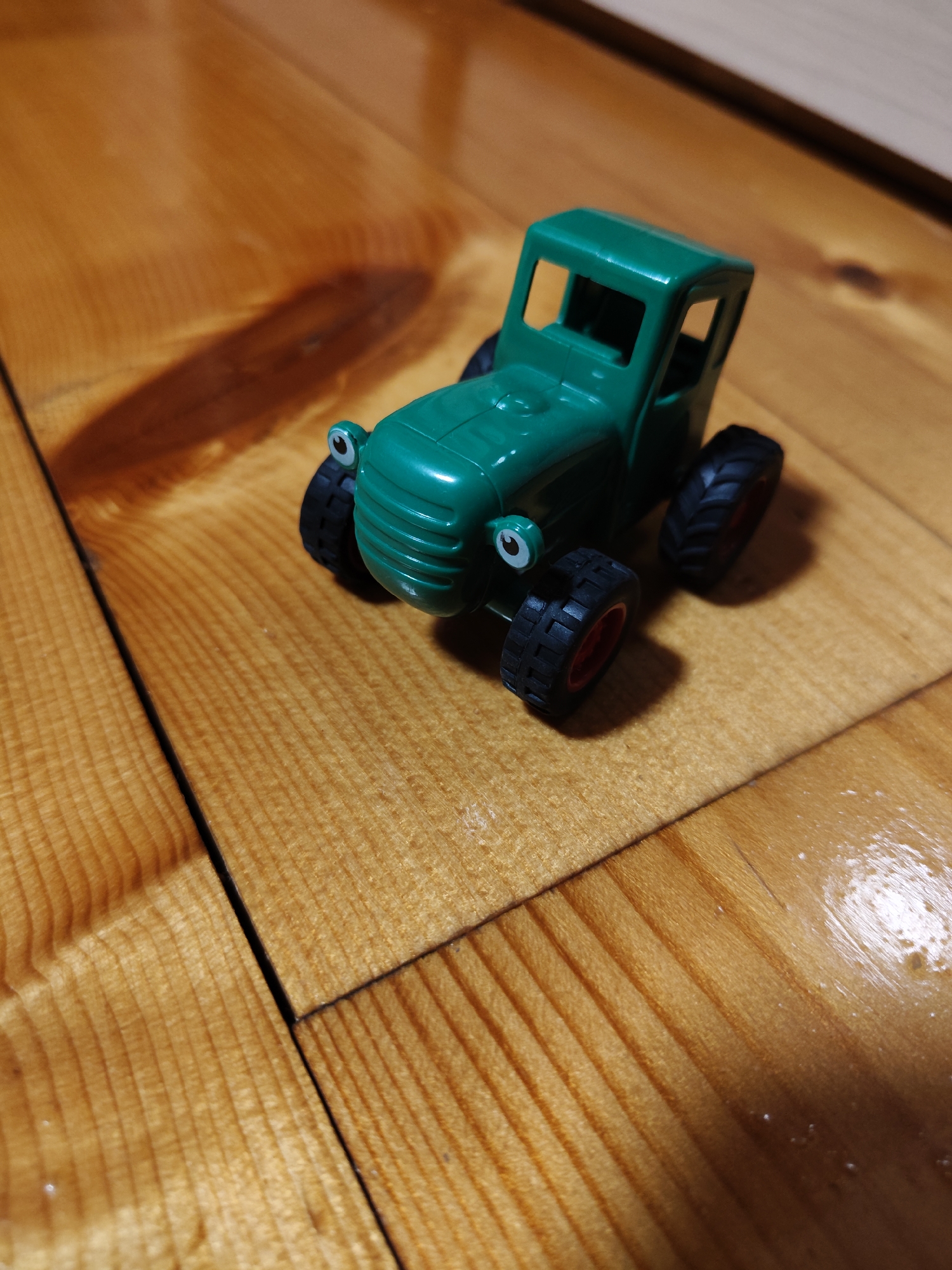 A green toy tractor sits on a wooden floor. The tractor is facing left and has large black wheels with red rims. It has a cab with open windows and a grille on the front. The wooden floor appears to be made of planks and has a warm, polished finish. There are some light reflections on the floor, suggesting it may be slightly wet or have a glossy coating. The background is out of focus but appears to be more of the wooden floor.